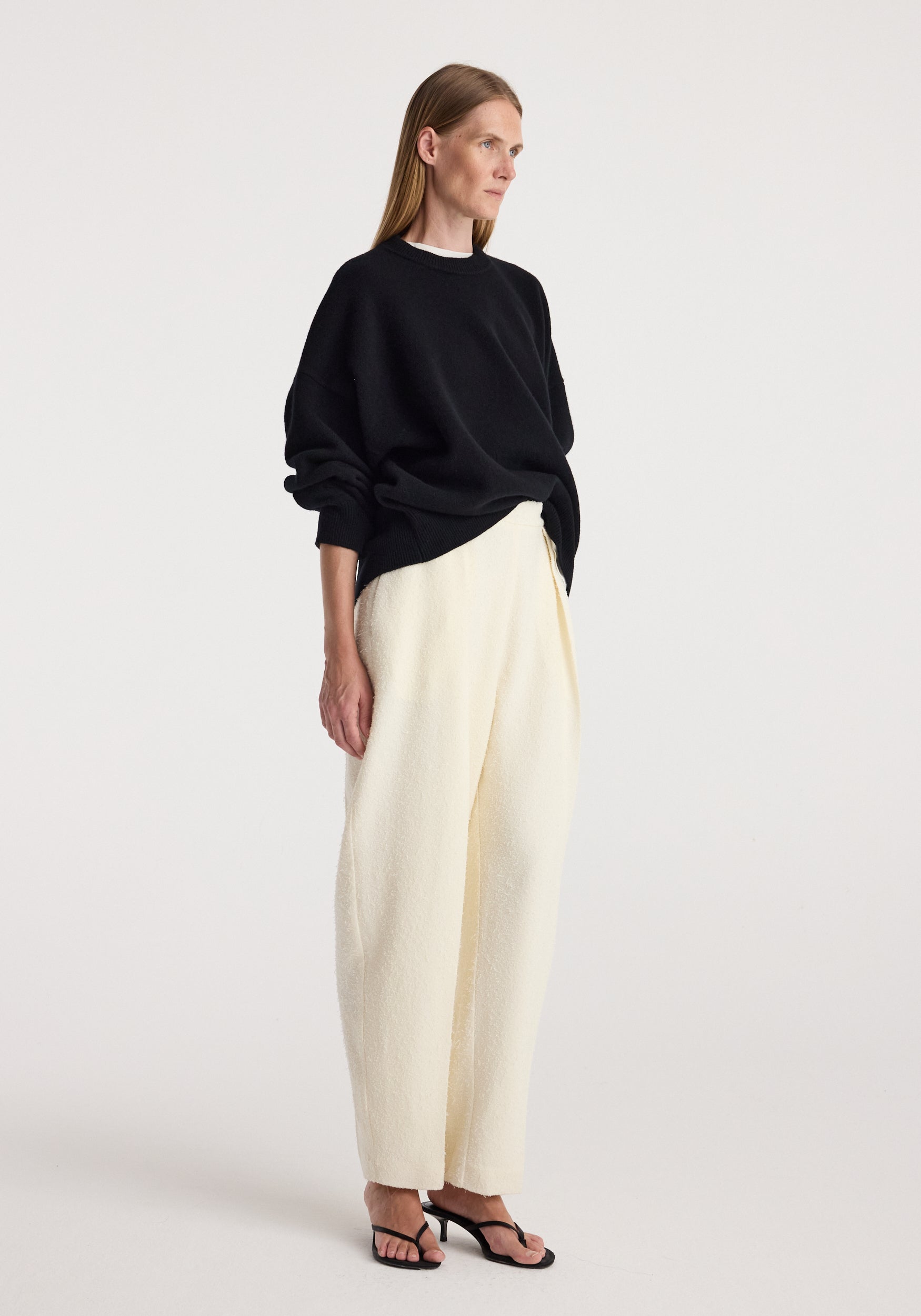 Woman wearing a loose-fitting Classic crewneck jumper | black paired with cream wide-leg pants and black open-toe heeled sandals, standing against a plain background.
