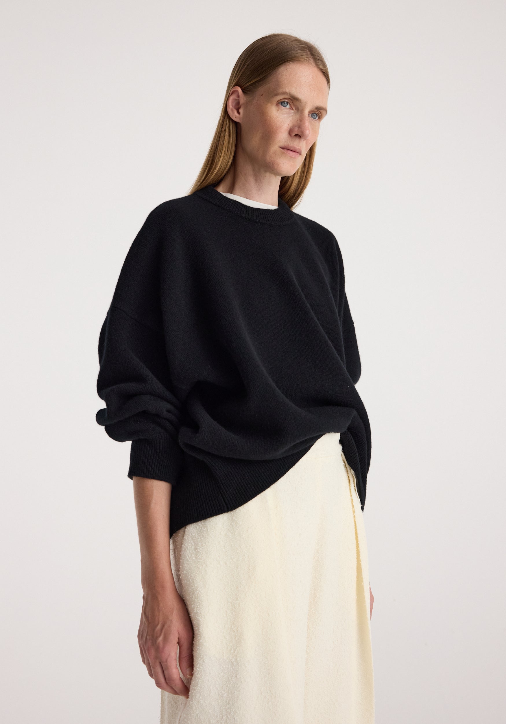 Woman wearing a black Classic crewneck jumper | black with long sleeves and ribbed cuffs, paired with a light-colored skirt, standing against a plain background.
