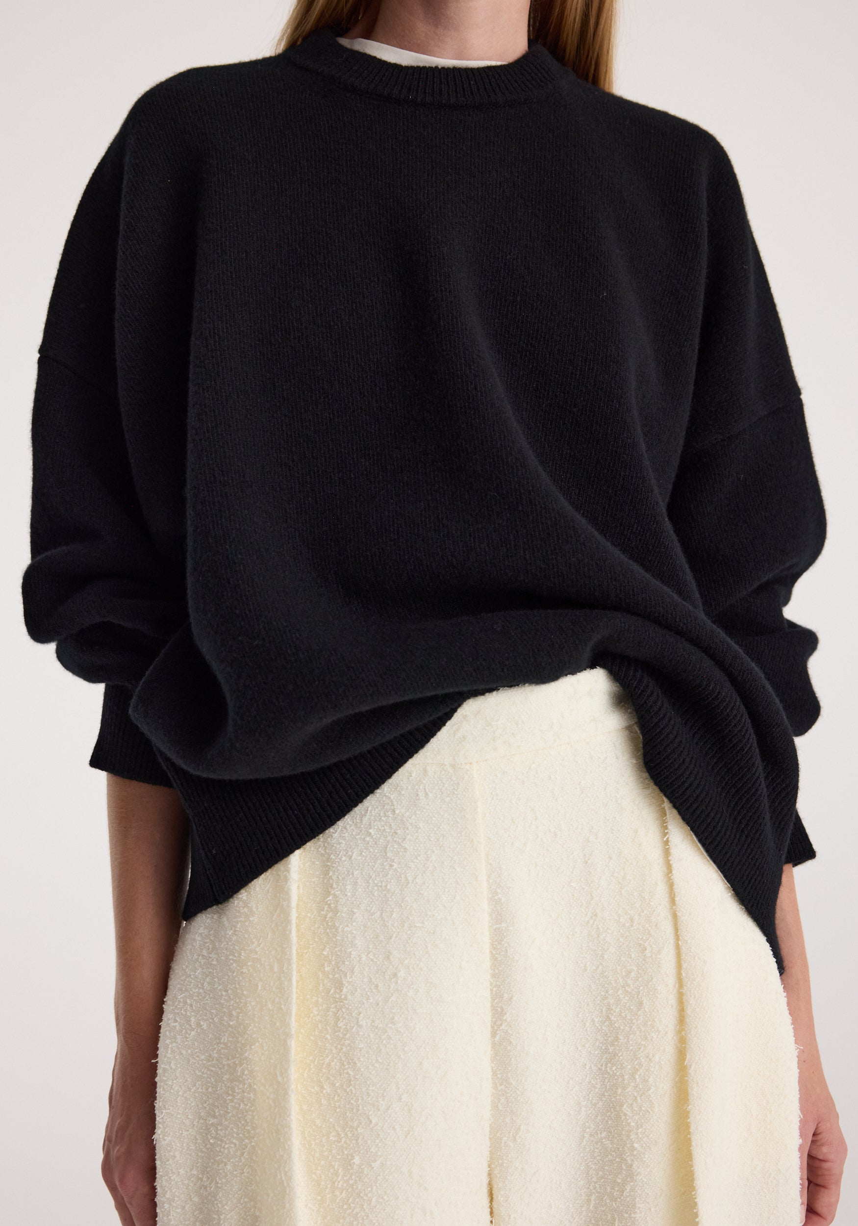 Close-up of a person wearing a Classic crewneck jumper | black with a relaxed fit over white textured pants, showcasing the soft knit fabric and casual style.