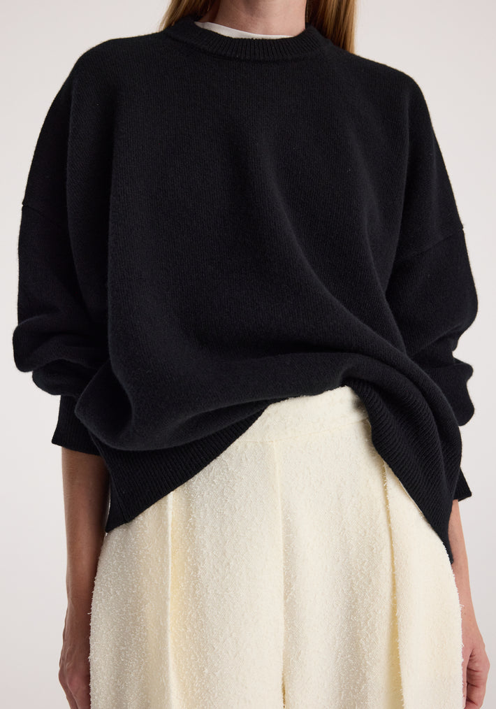Close-up of a person wearing a Classic crewneck jumper | black with a relaxed fit over white textured pants, showcasing the soft knit fabric and casual style.