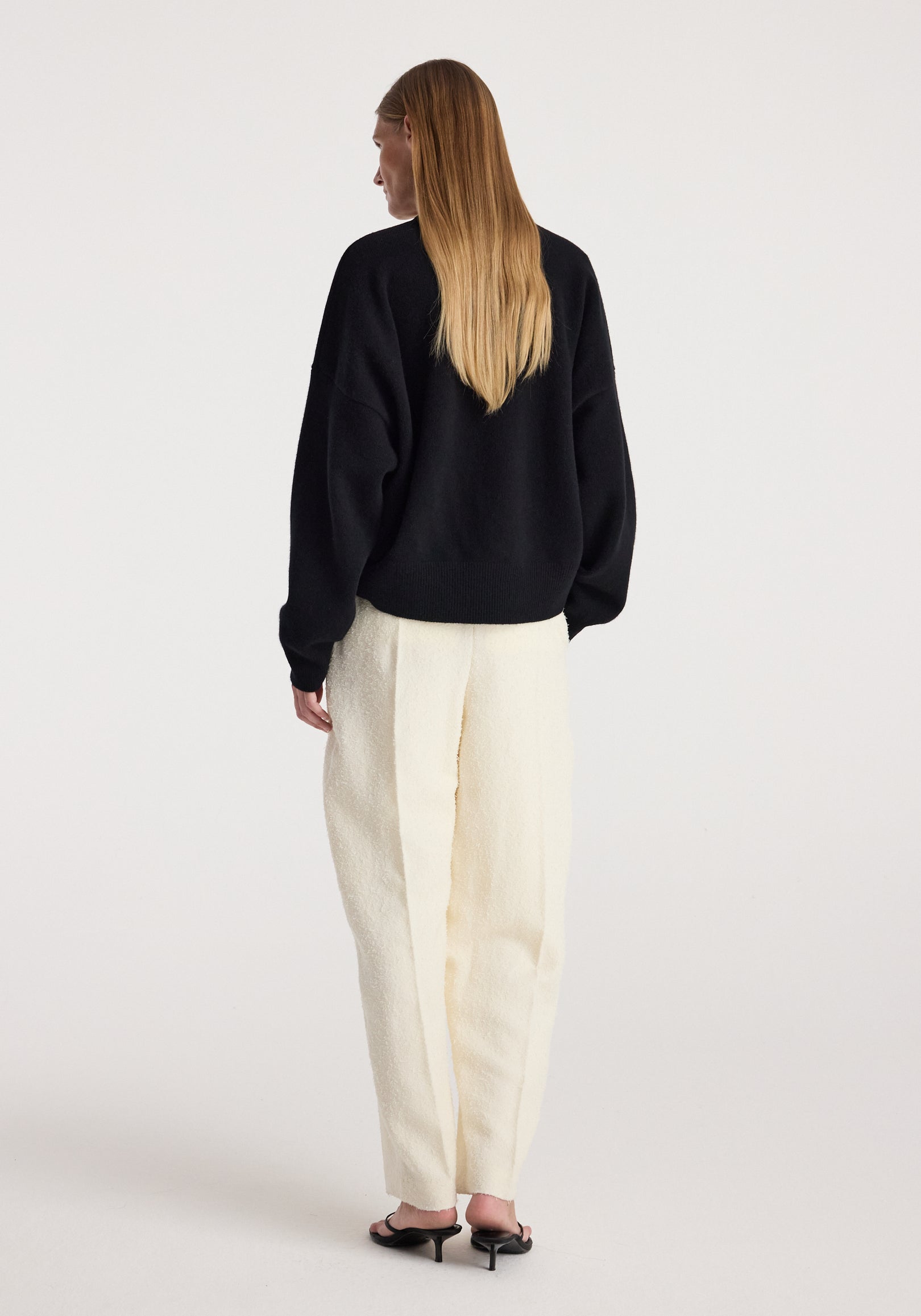 Woman seen from behind wearing a Classic crewneck jumper | black paired with loose cream-colored pants and black heeled sandals against a plain background.
