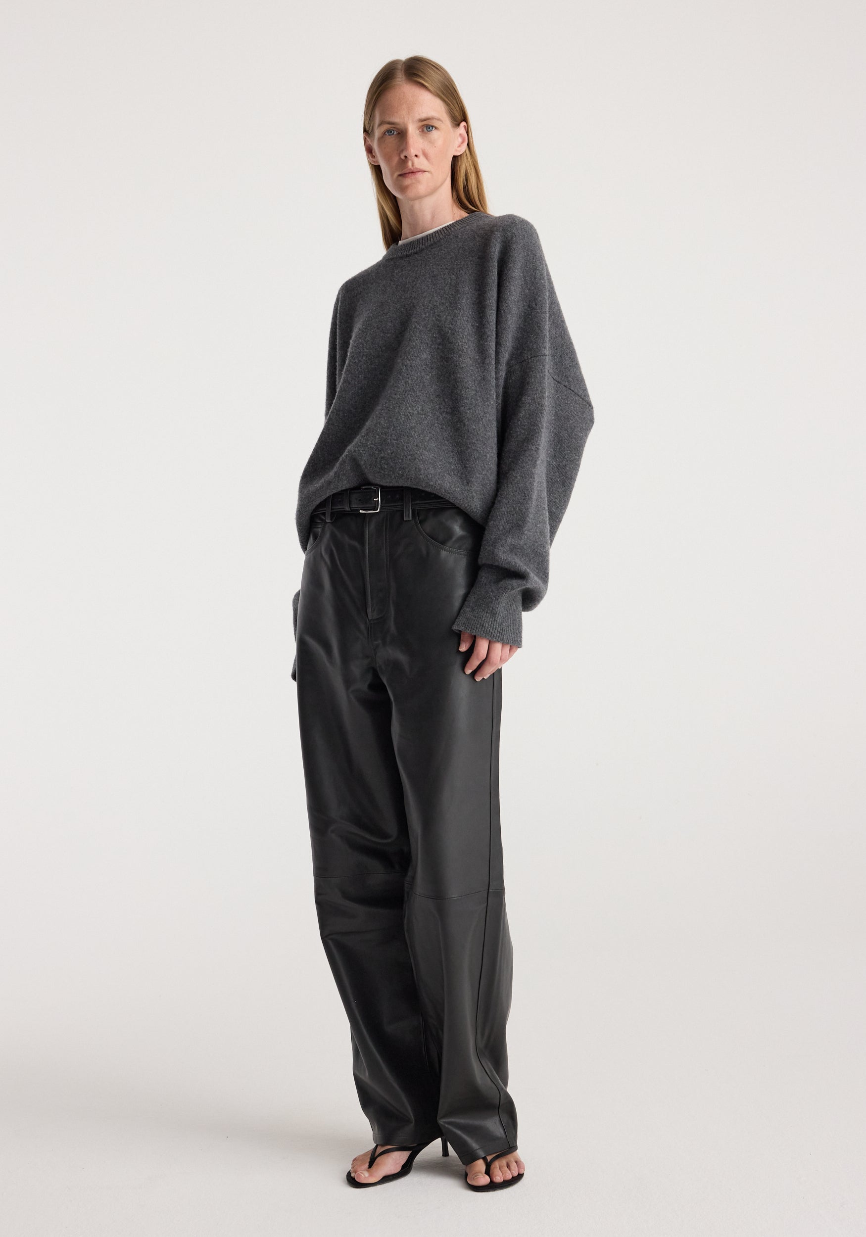 Woman wearing a relaxed fit Classic crewneck jumper | dark grey melange paired with black high-waisted pants and black open-toe heeled sandals against a plain background.