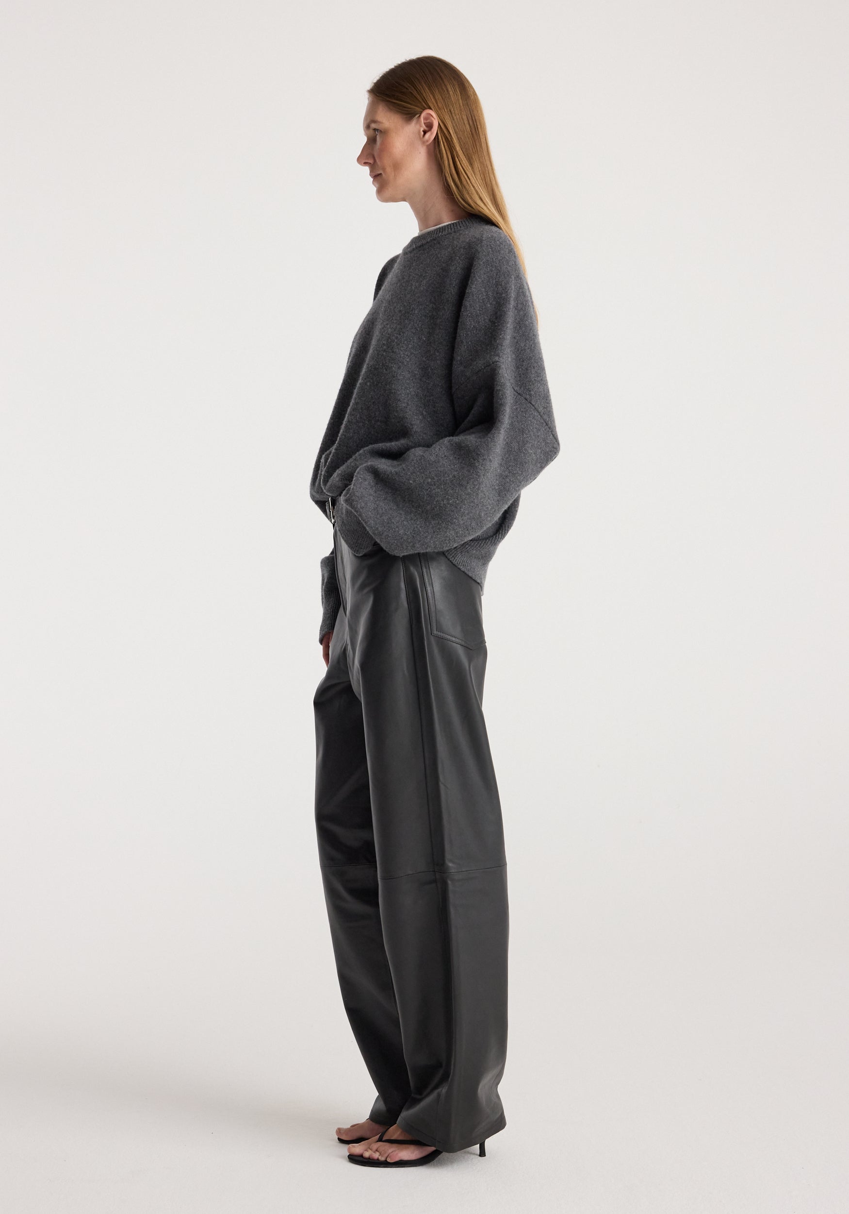 Woman wearing a dark grey melange classic crewneck jumper paired with black wide-leg leather pants and black heeled sandals, standing in profile against a plain background.