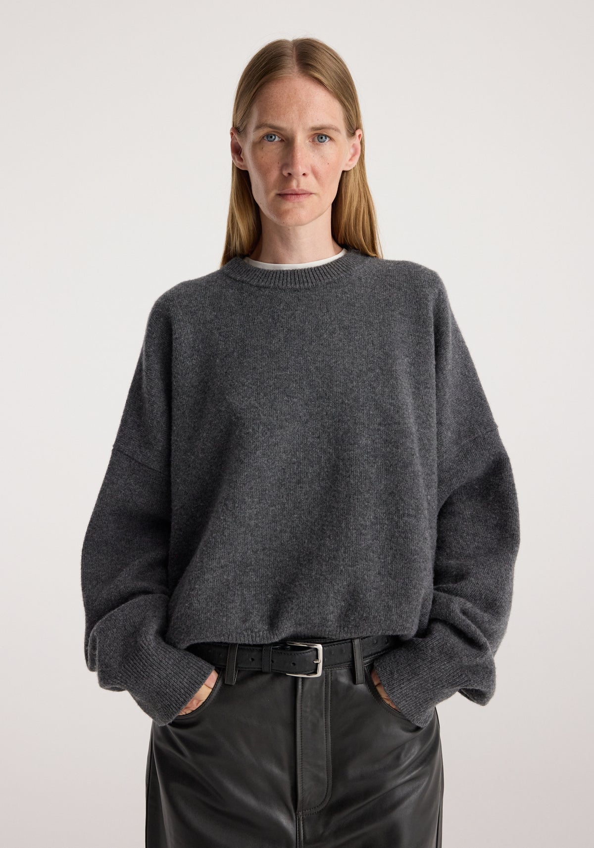 Woman wearing a Classic crewneck jumper | dark grey melange with a relaxed fit and soft texture, styled with black leather pants and a belt in a neutral background setting.