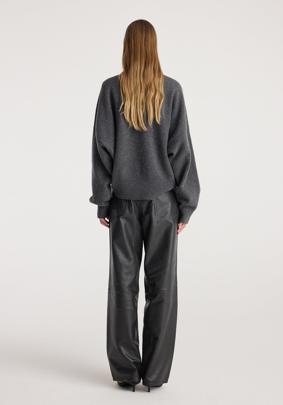 Woman wearing a dark grey melange Classic crewneck jumper with long sleeves, paired with black leather pants, viewed from the back on a plain background.
