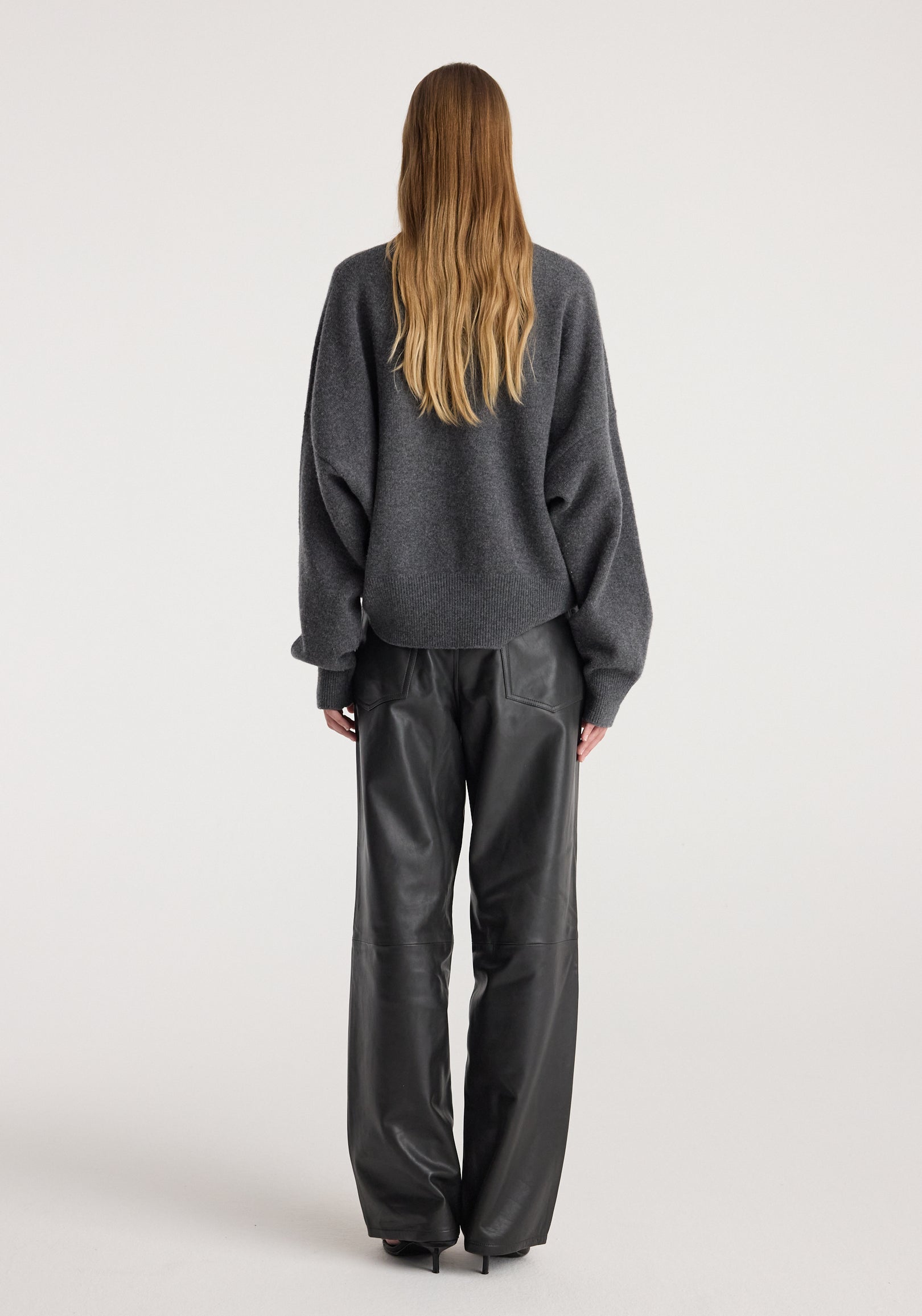 Woman wearing a dark grey melange Classic crewneck jumper with long sleeves, paired with black leather pants, viewed from the back on a plain background.