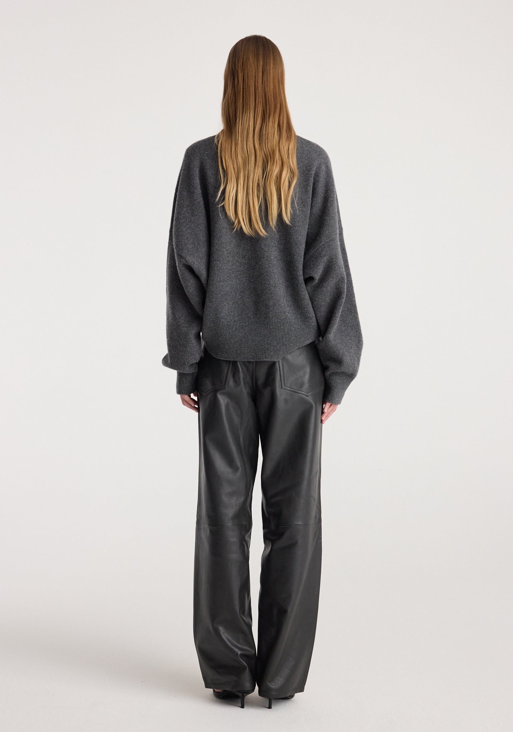 Woman wearing a dark grey melange Classic crewneck jumper with long sleeves, paired with black leather pants, viewed from the back on a plain background.