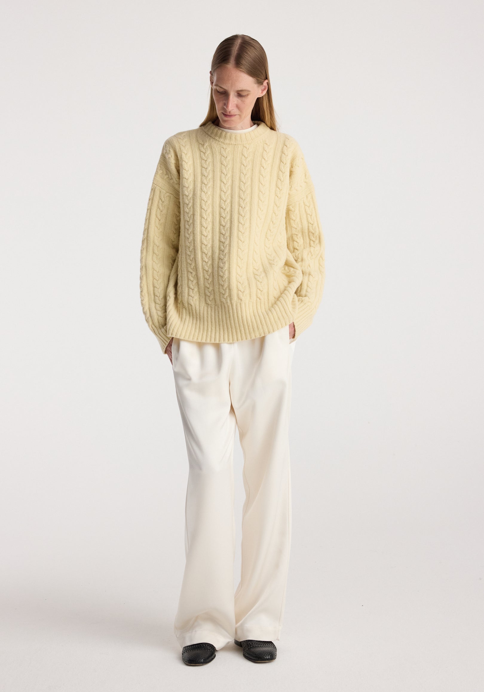 Woman wearing a cream-colored cable knit jumper | butter with long sleeves, paired with loose white trousers and black shoes, standing against a plain white background.