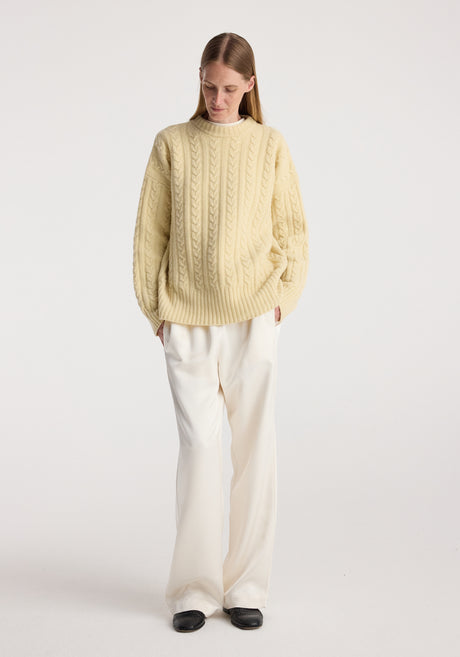 Woman wearing a cream-colored cable knit jumper | butter with long sleeves, paired with loose white trousers and black shoes, standing against a plain white background.