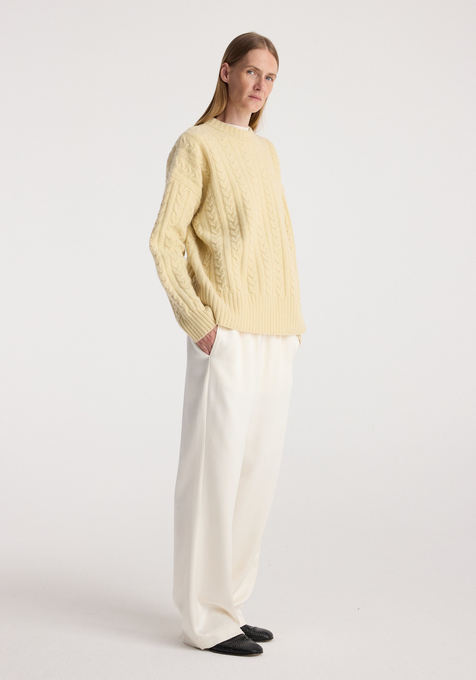 Woman wearing a cream yellow cable knit jumper | butter paired with loose white trousers and black shoes, standing against a plain white background.