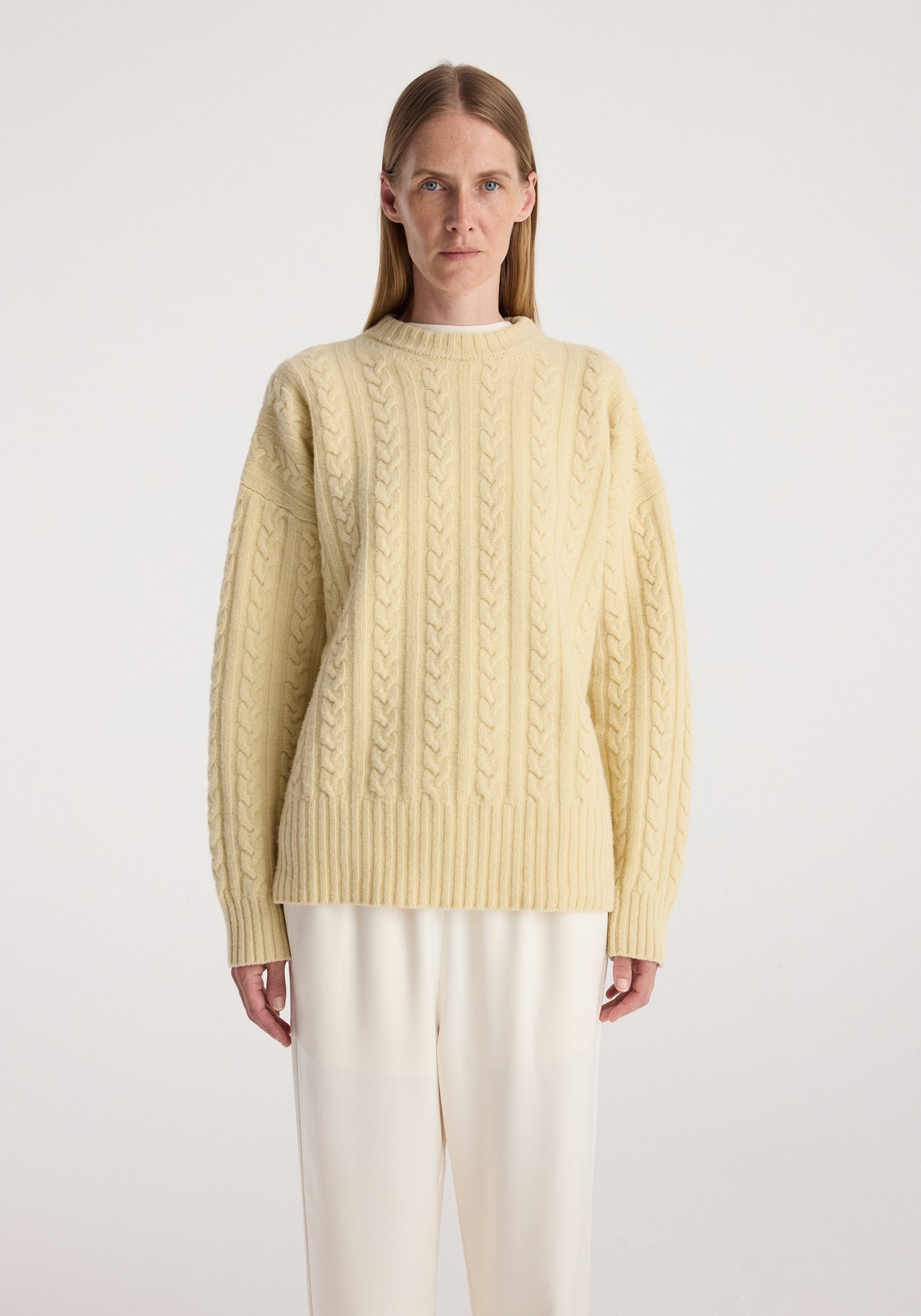 Woman wearing a butter-colored cable knit jumper | butter with a textured pattern, long sleeves, and ribbed cuffs, paired with white pants against a plain background.