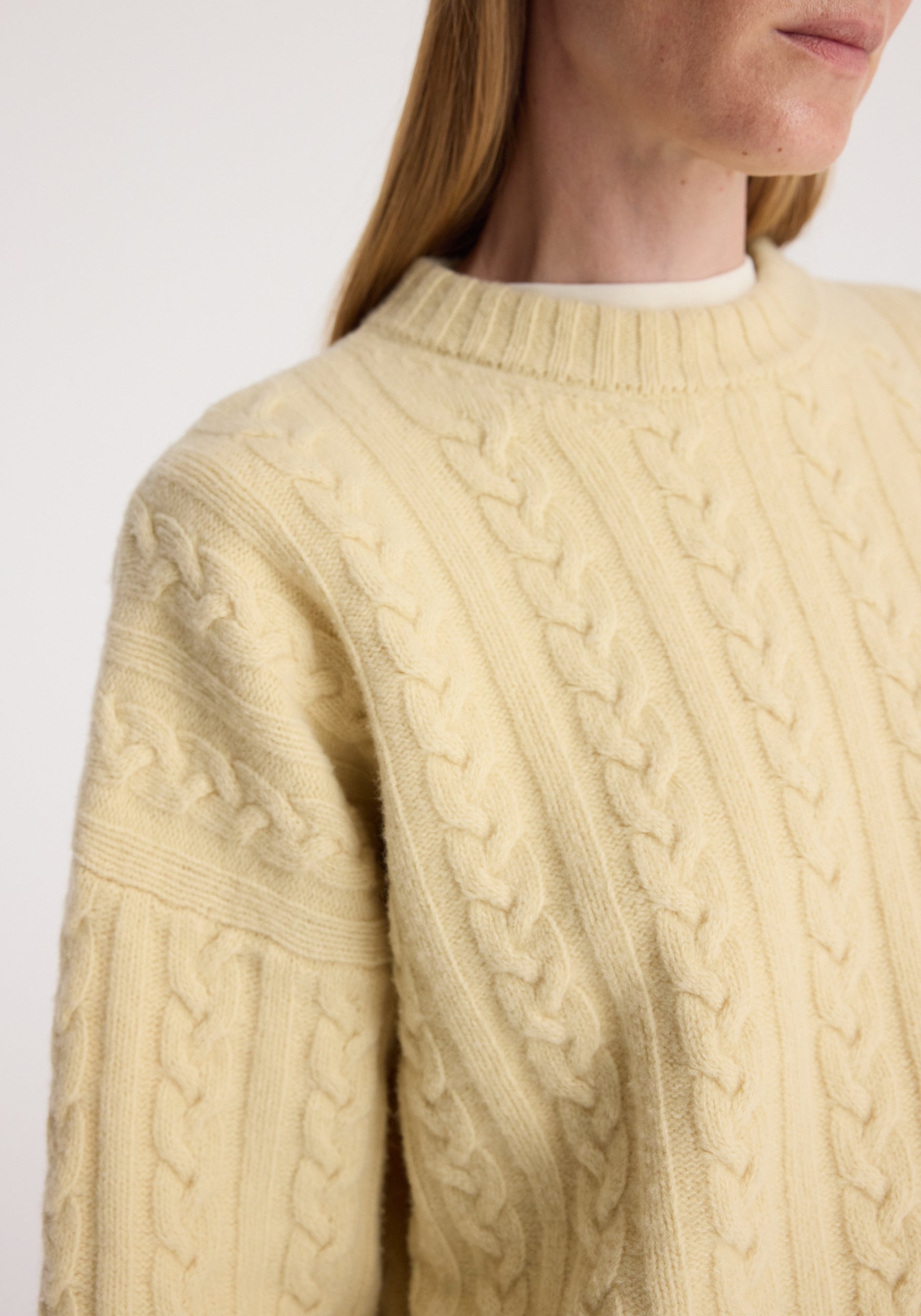 Close-up of a person wearing a soft, textured cream-colored Cable knit jumper | butter with a ribbed crew neckline and detailed braided patterns.