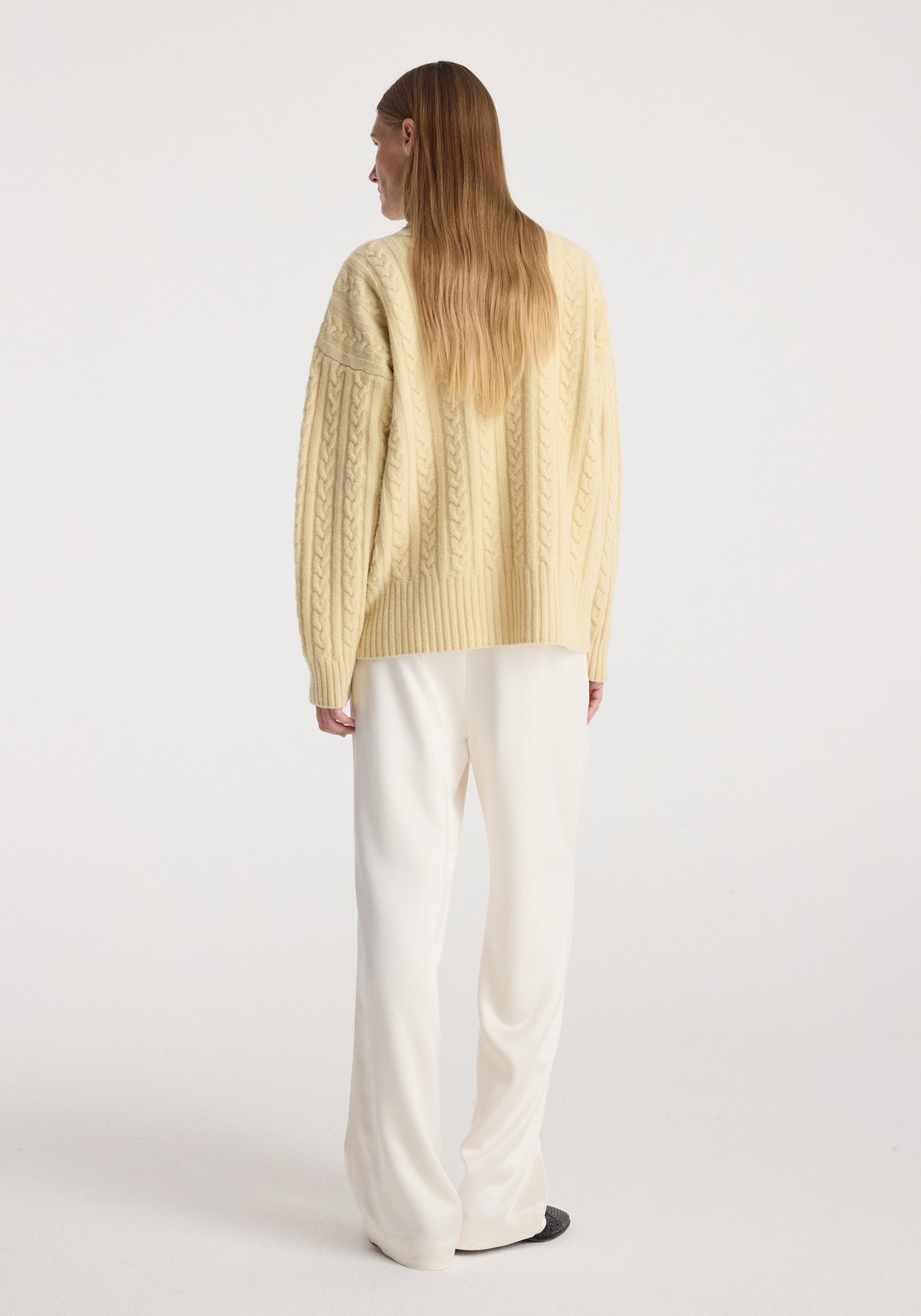 Woman wearing a pale yellow Cable knit jumper | butter with long sleeves and intricate cable patterns, paired with loose white pants, standing facing away on a plain background.