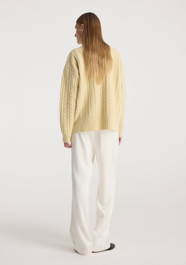 Woman wearing a pale yellow Cable knit jumper | butter with long sleeves and intricate cable patterns, paired with loose white pants, standing facing away on a plain background.