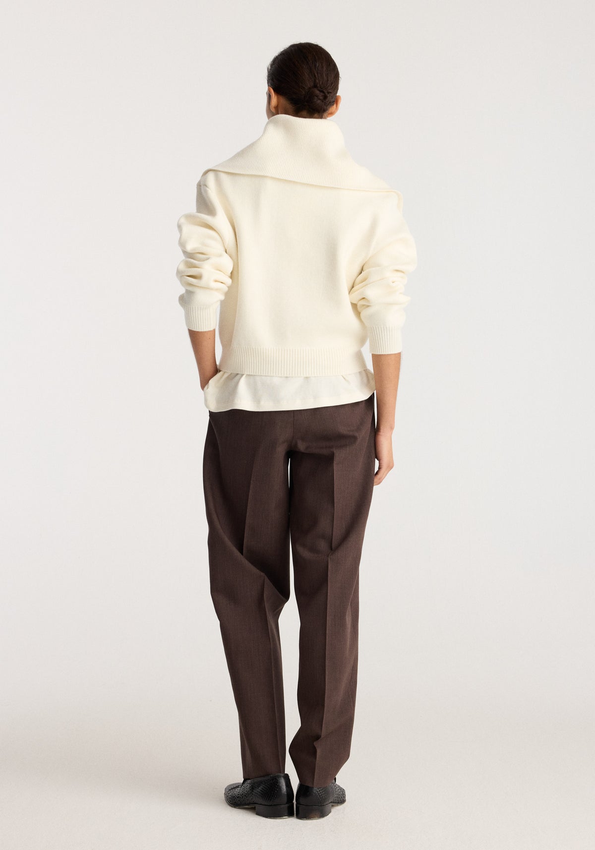 Back view of a model wearing the Reversible sailor zip cardigan | cream with sleeves pushed up and dark brown trousers in a neutral studio setting