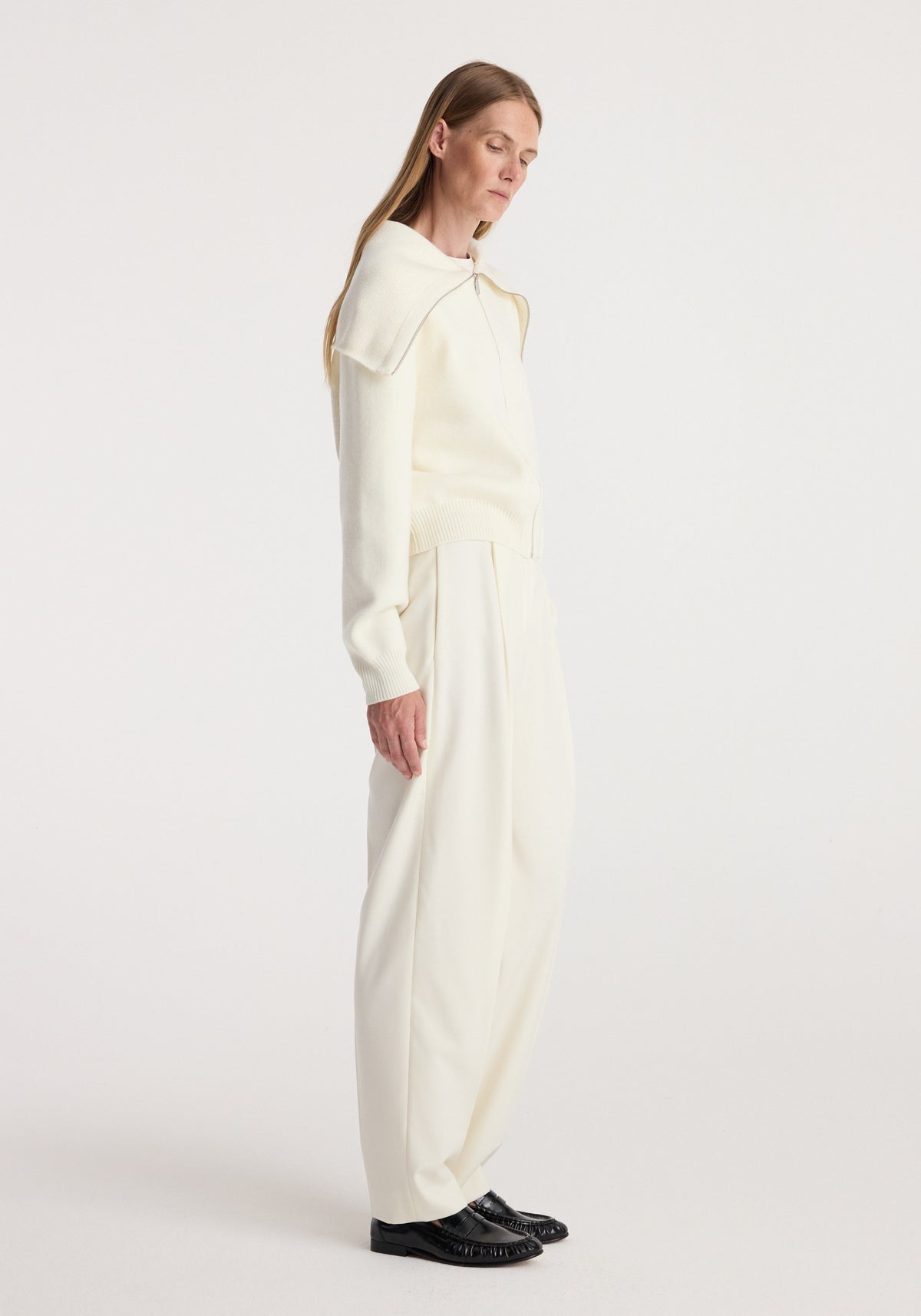 Woman wearing a cream long-sleeve Two way sailor zip cardigan | cream paired with loose cream pants and black loafers, standing in profile against a plain background.
