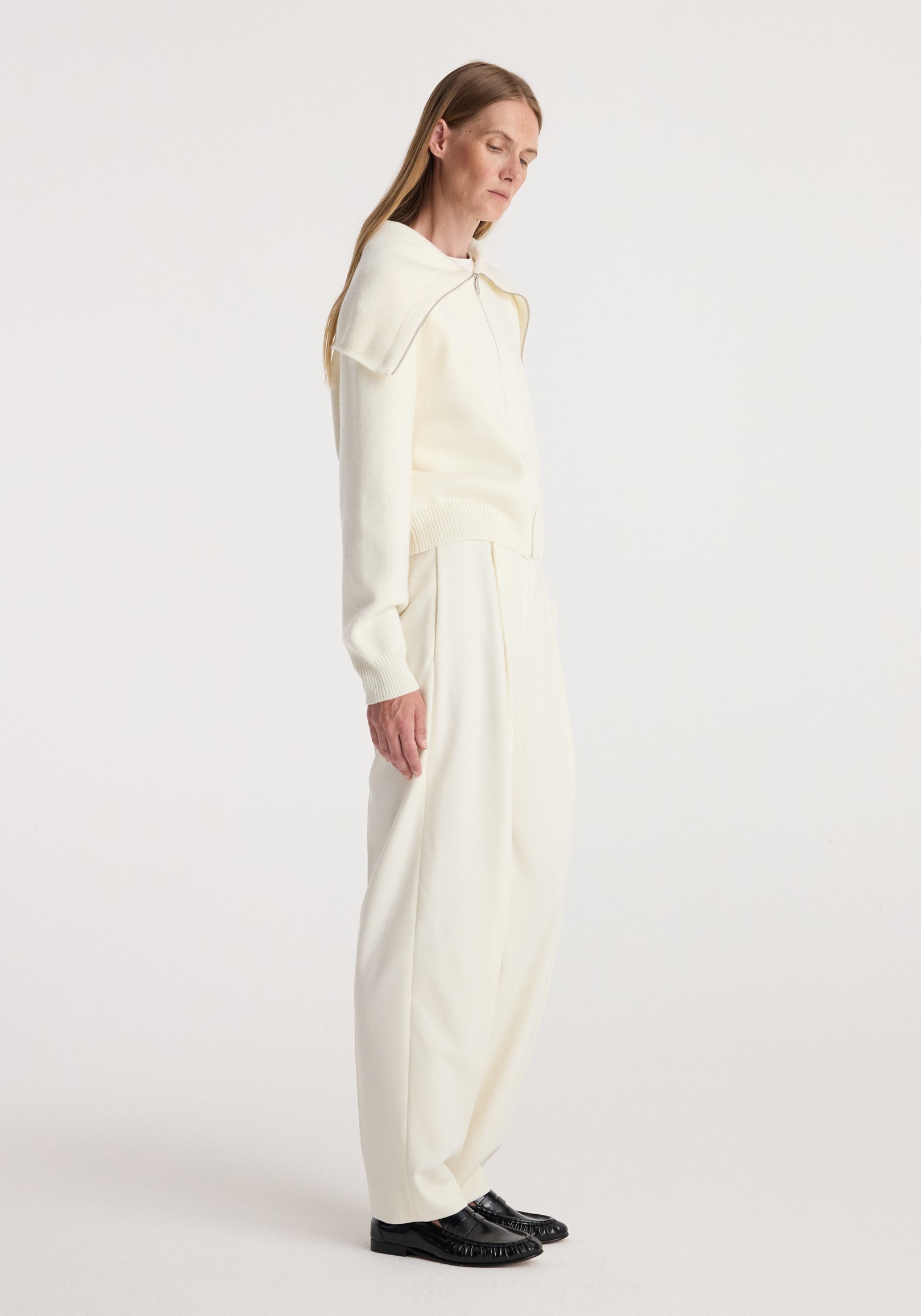 Woman wearing a cream long-sleeve Two way sailor zip cardigan | cream paired with loose cream pants and black loafers, standing in profile against a plain background.
