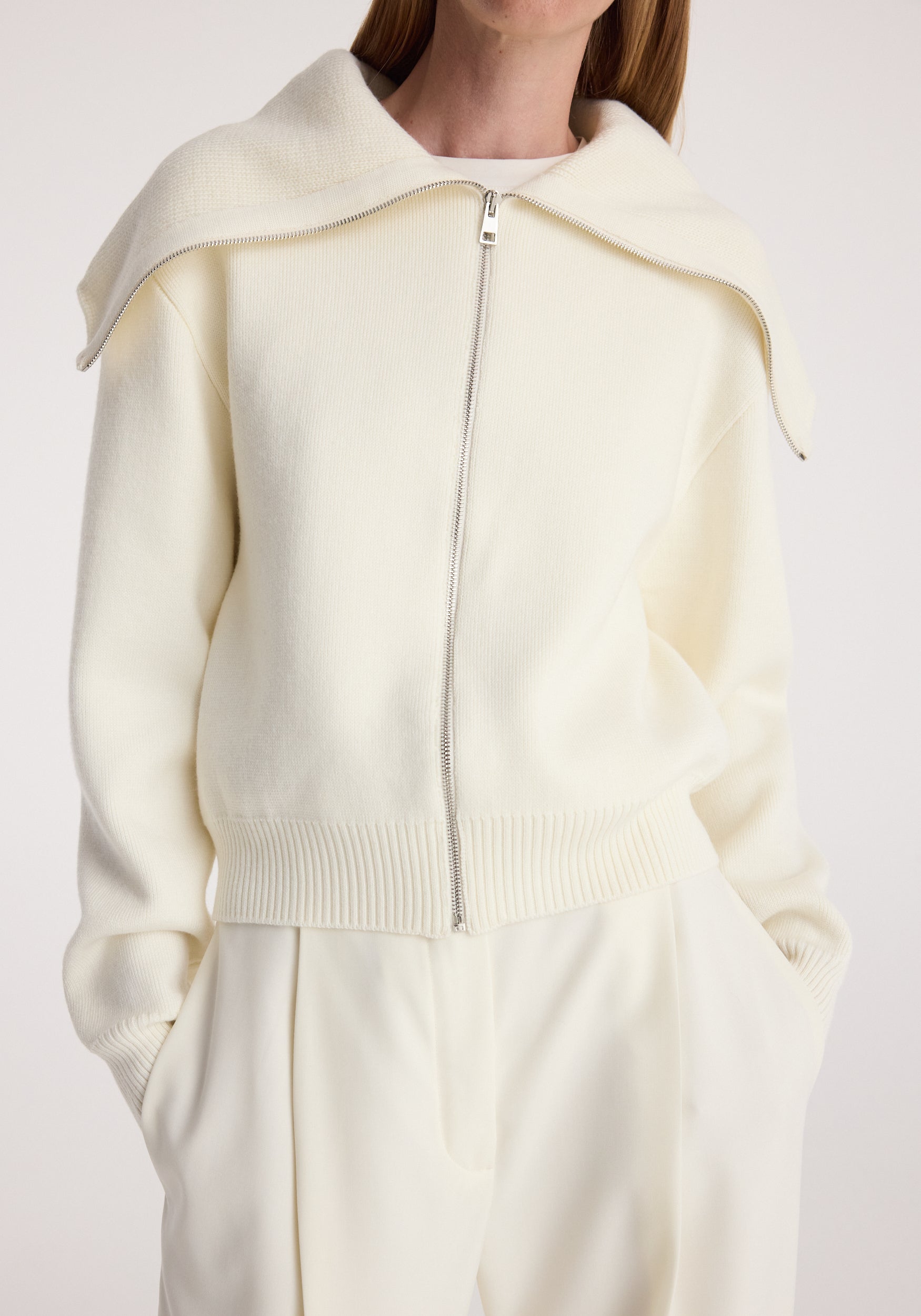 Woman wearing a cream two way sailor zip cardigan | cream with a unique wide collar and ribbed cuffs, paired with matching cream pants against a plain background.