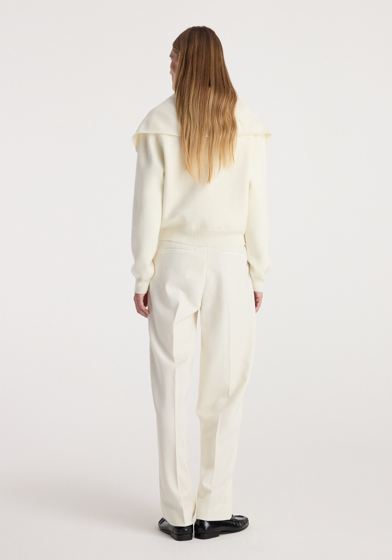 Woman wearing cream two way sailor zip cardigan | cream with long sleeves and collar, paired with matching cream trousers and black shoes, standing with back facing camera.