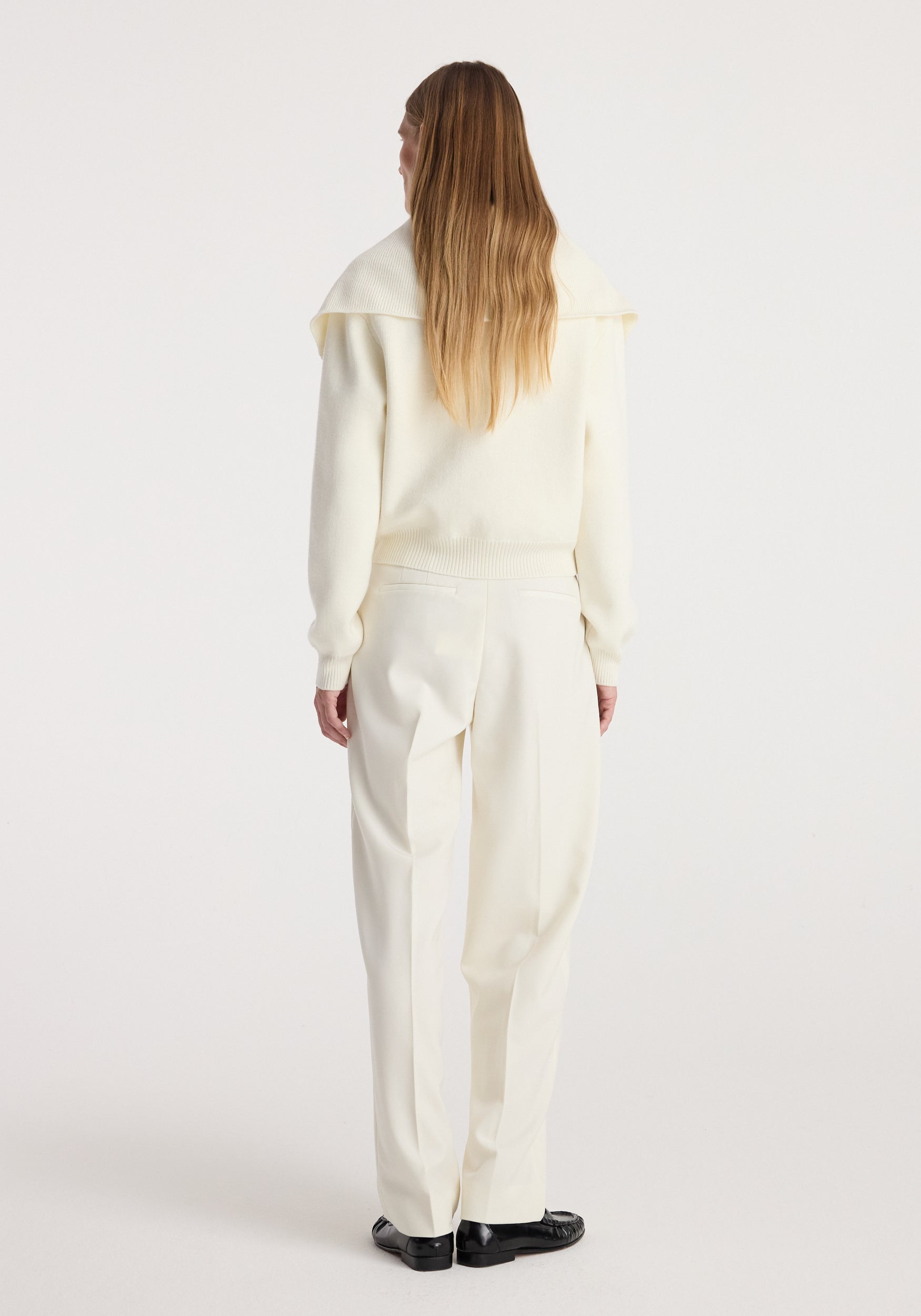 Woman wearing cream two way sailor zip cardigan | cream with long sleeves and collar, paired with matching cream trousers and black shoes, standing with back facing camera.
