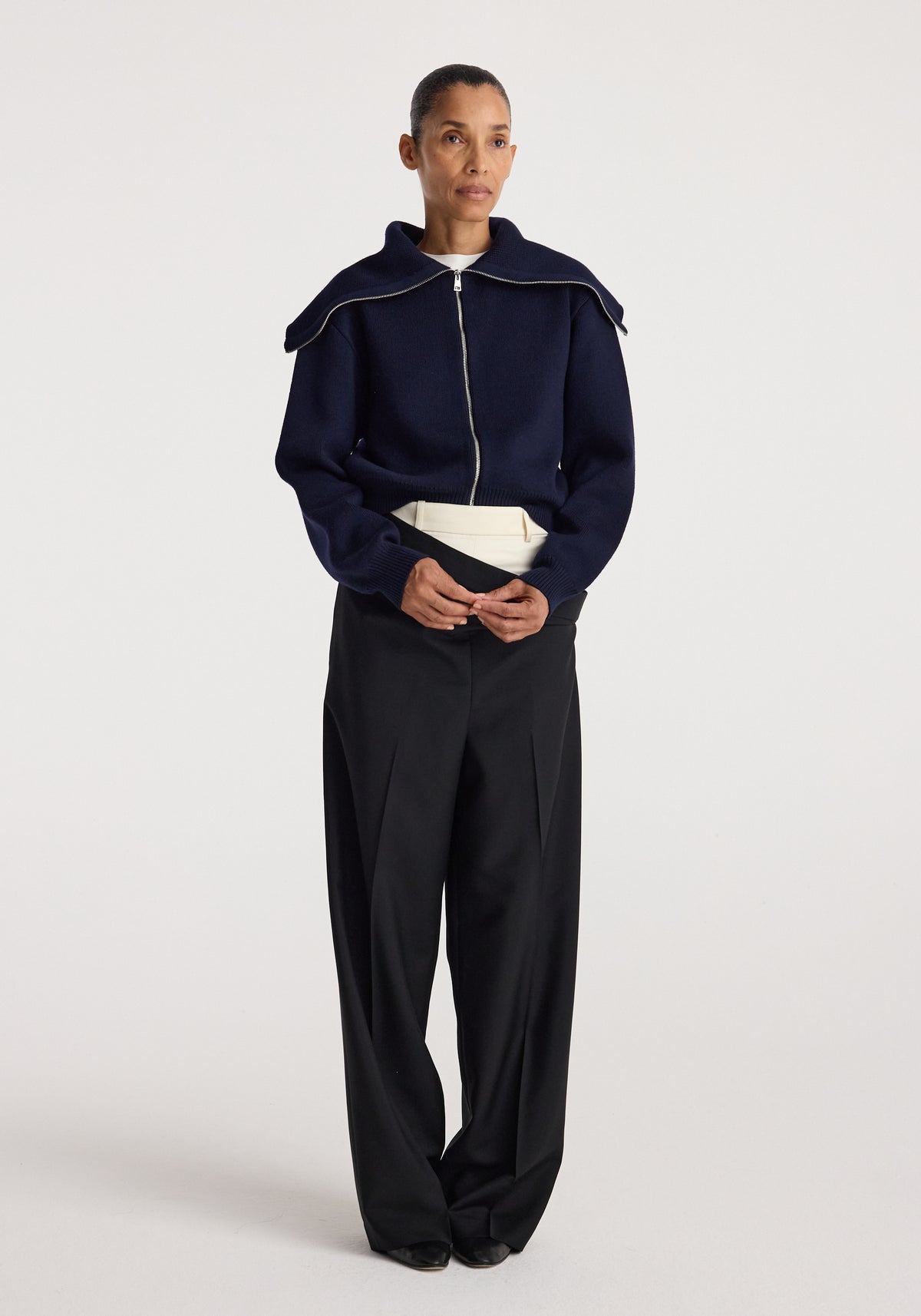 Woman wearing a navy two way sailor zip cardigan | navy with a large sailor collar zipped up, paired with high-waisted black wide-leg pants and a white belt.