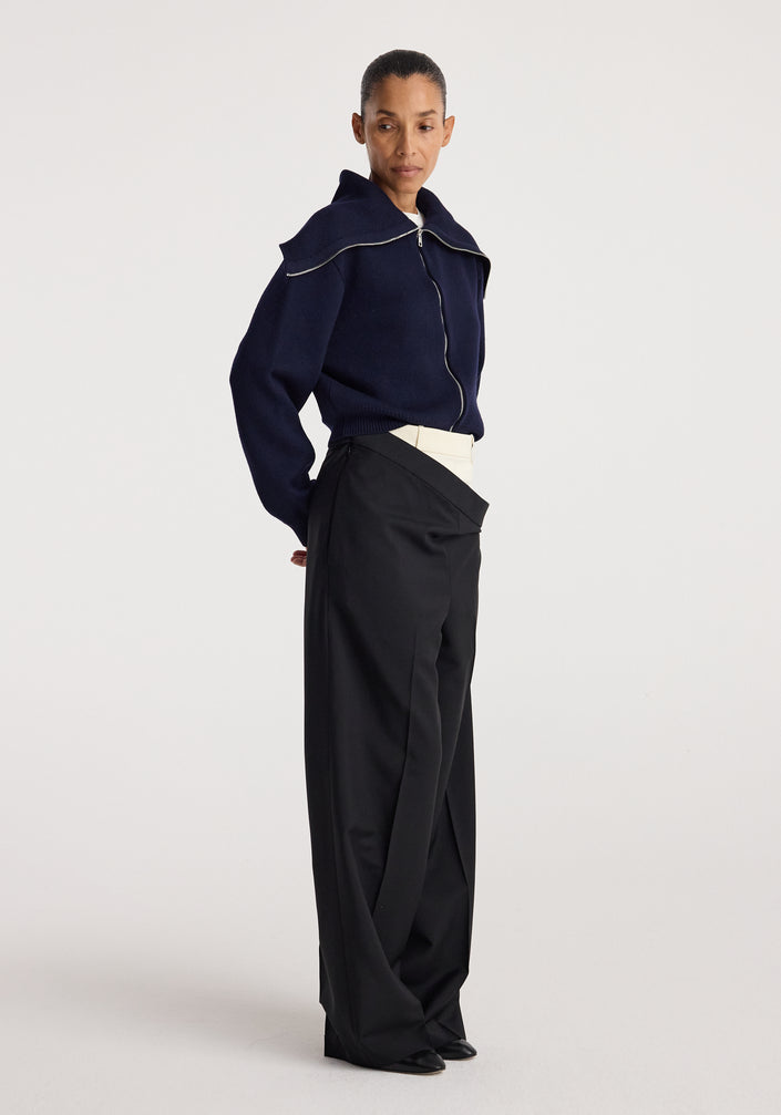 Woman wearing a navy two way sailor zip cardigan | navy with a large sailor collar zipped up, paired with high-waisted black wide-leg pants and a white belt.