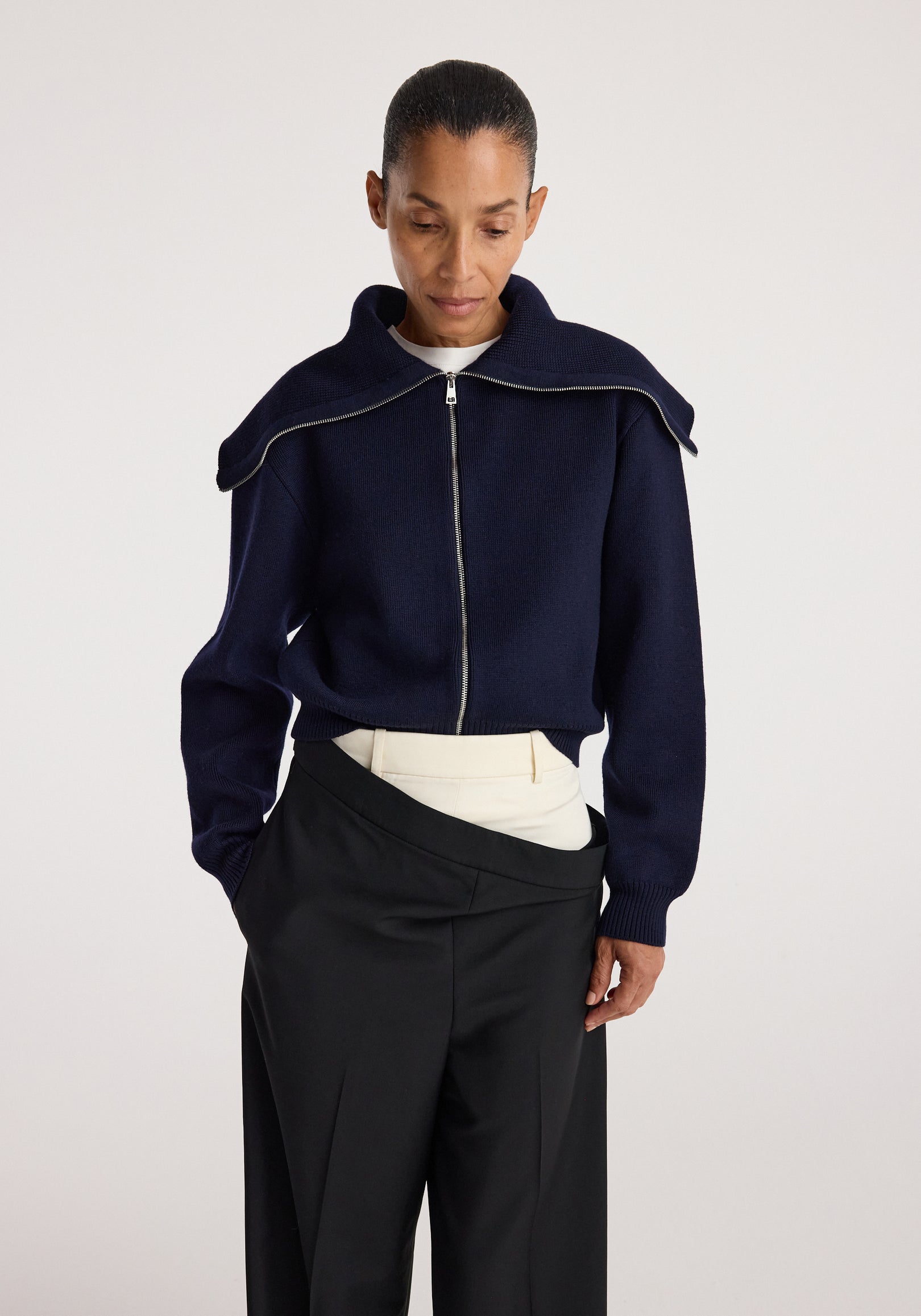 Woman wearing a navy two way sailor zip cardigan | navy with a wide collar, paired with layered light and dark trousers against a plain background.