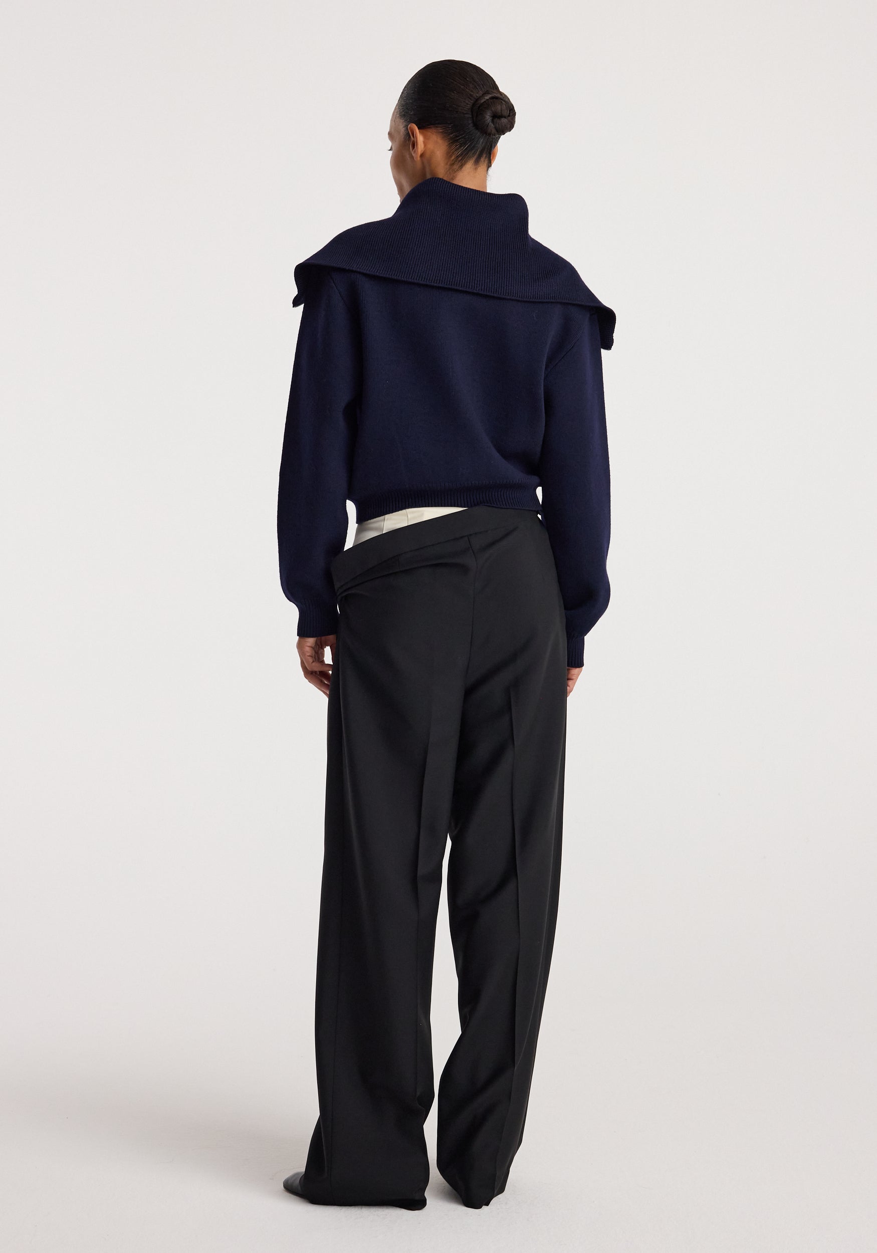 Woman seen from behind wearing a navy blue Two way sailor zip cardigan | navy with a wide collar paired with black wide-leg trousers against a plain background.