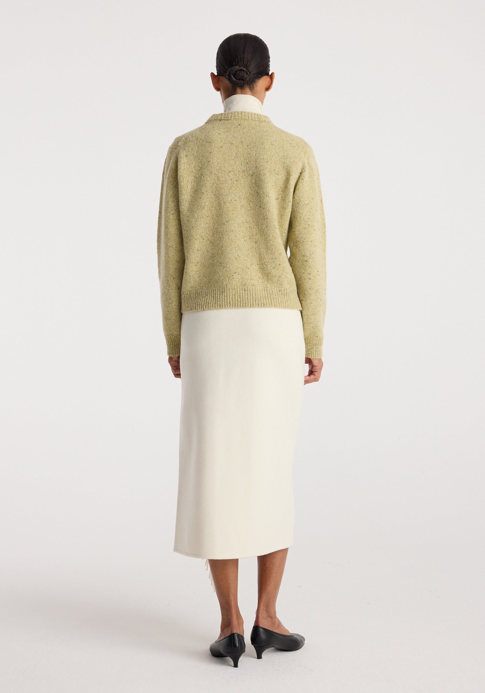 Woman wearing a fitted mouliné crewneck | butter mouliné sweater shown from the back, paired with a long white skirt and black low-heeled shoes in a minimalist studio setting.