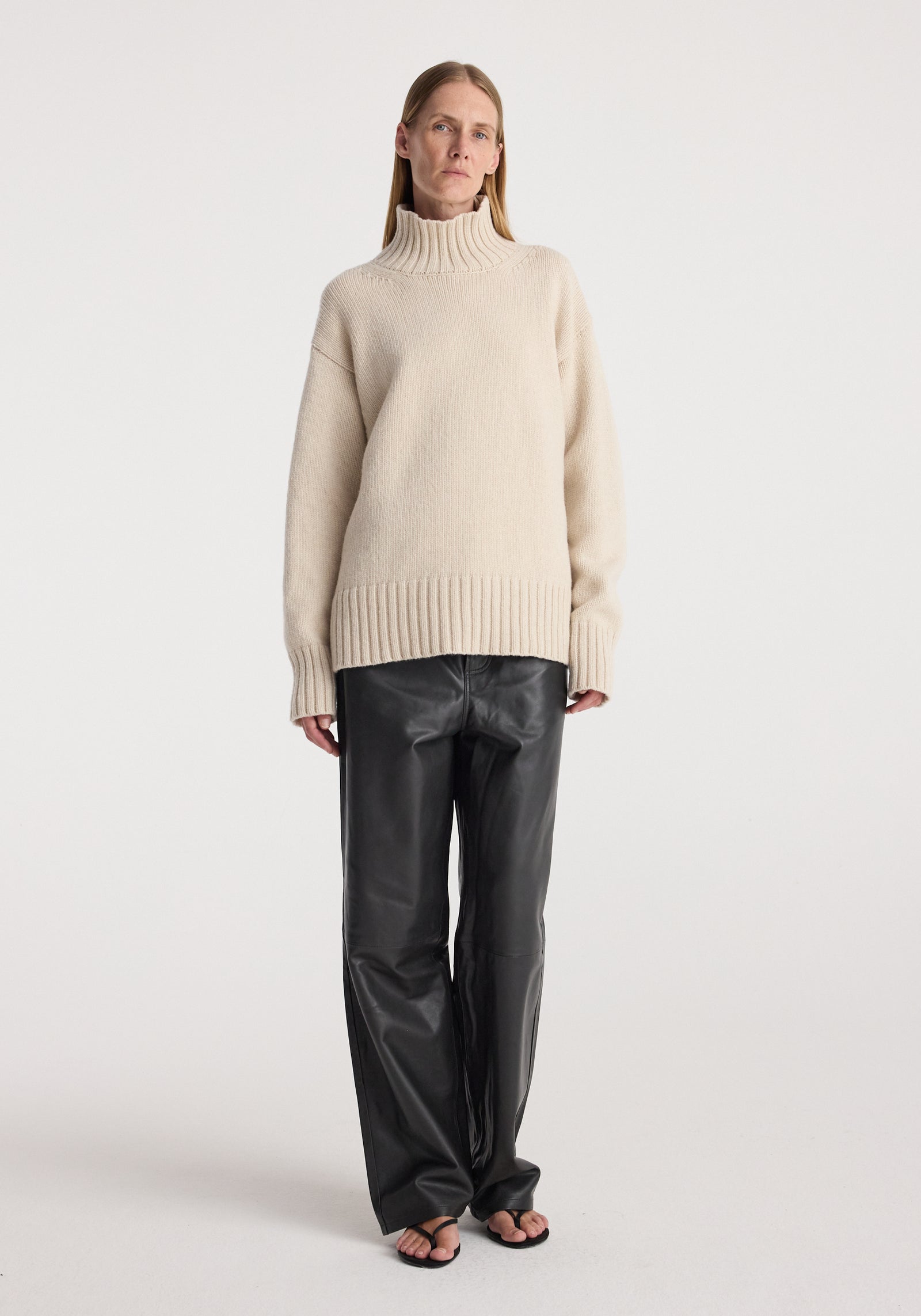 Woman wearing a wool-cashmere turtleneck | oat sweater paired with black leather pants and black sandals, standing against a plain white background.