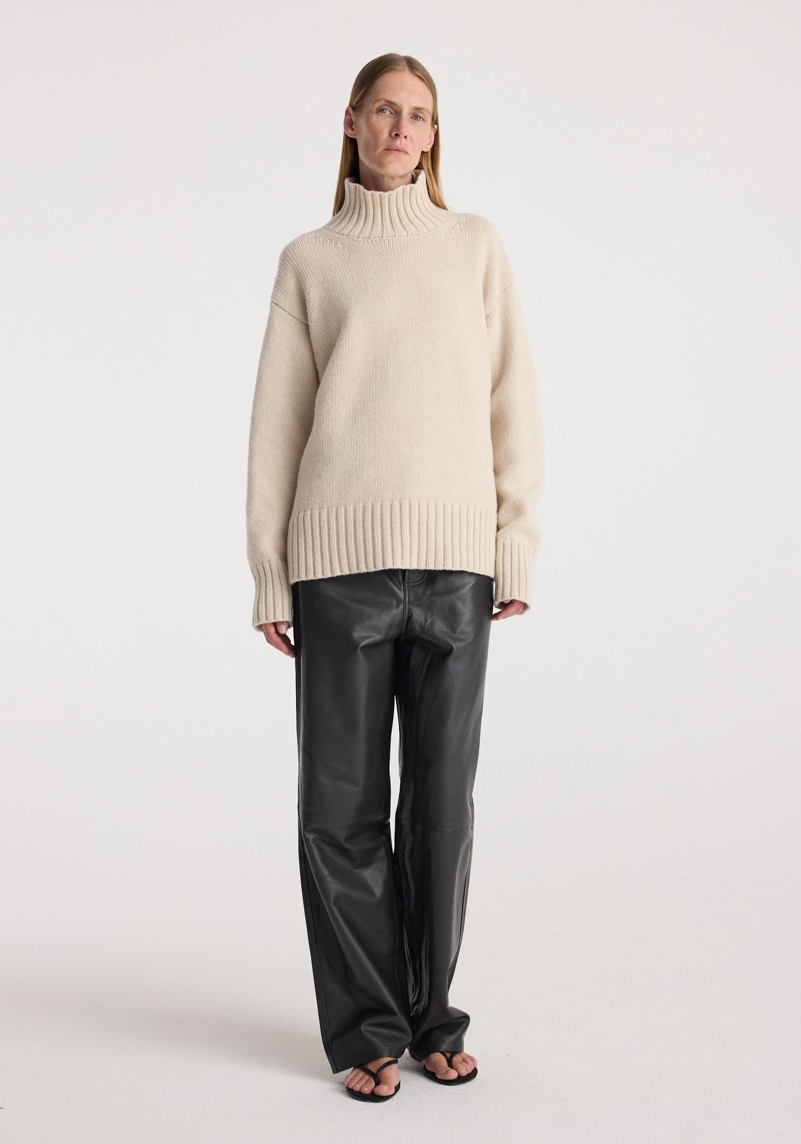 Woman wearing a wool-cashmere turtleneck | oat sweater paired with black leather pants and black sandals, standing against a plain white background.