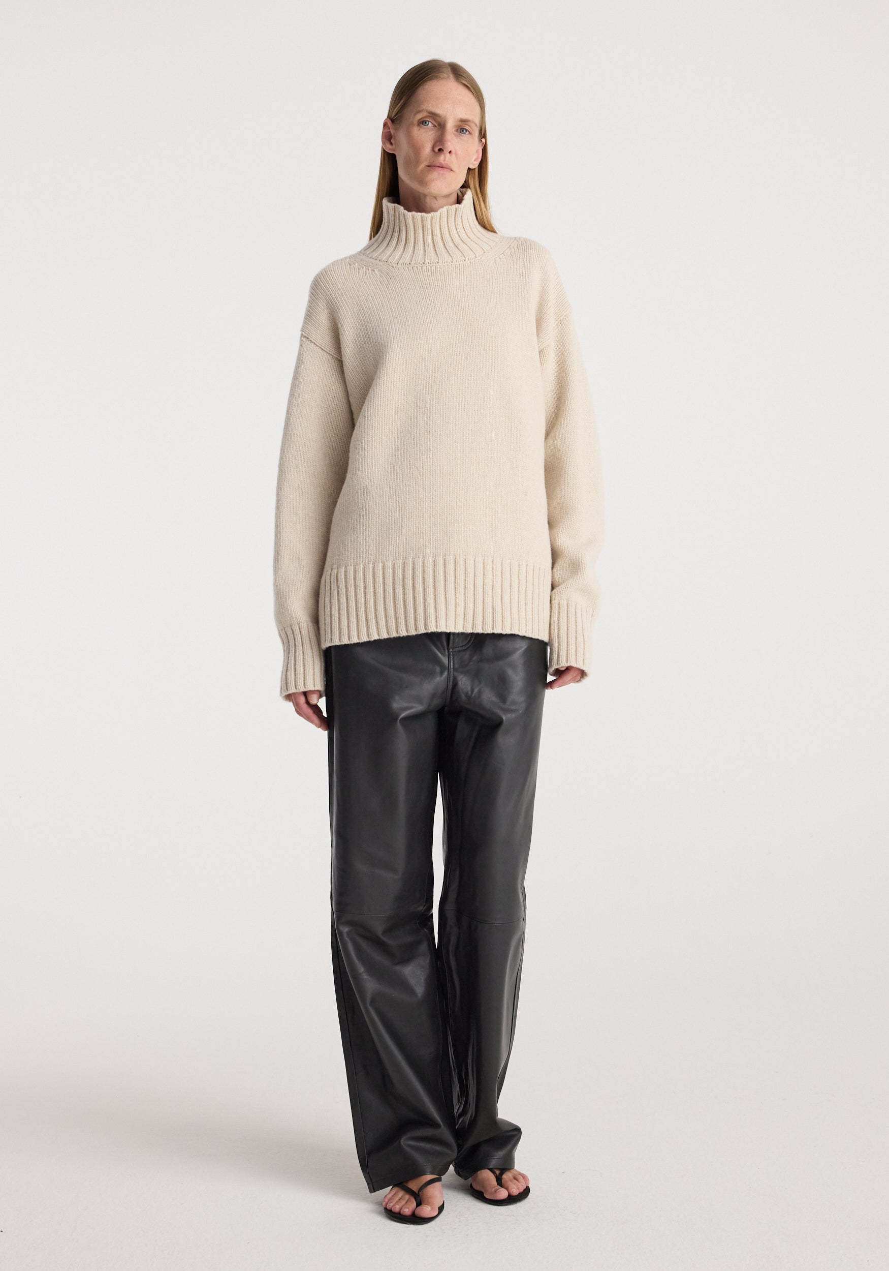 Woman wearing a wool-cashmere turtleneck | oat sweater paired with black leather pants and black sandals, standing against a plain white background.