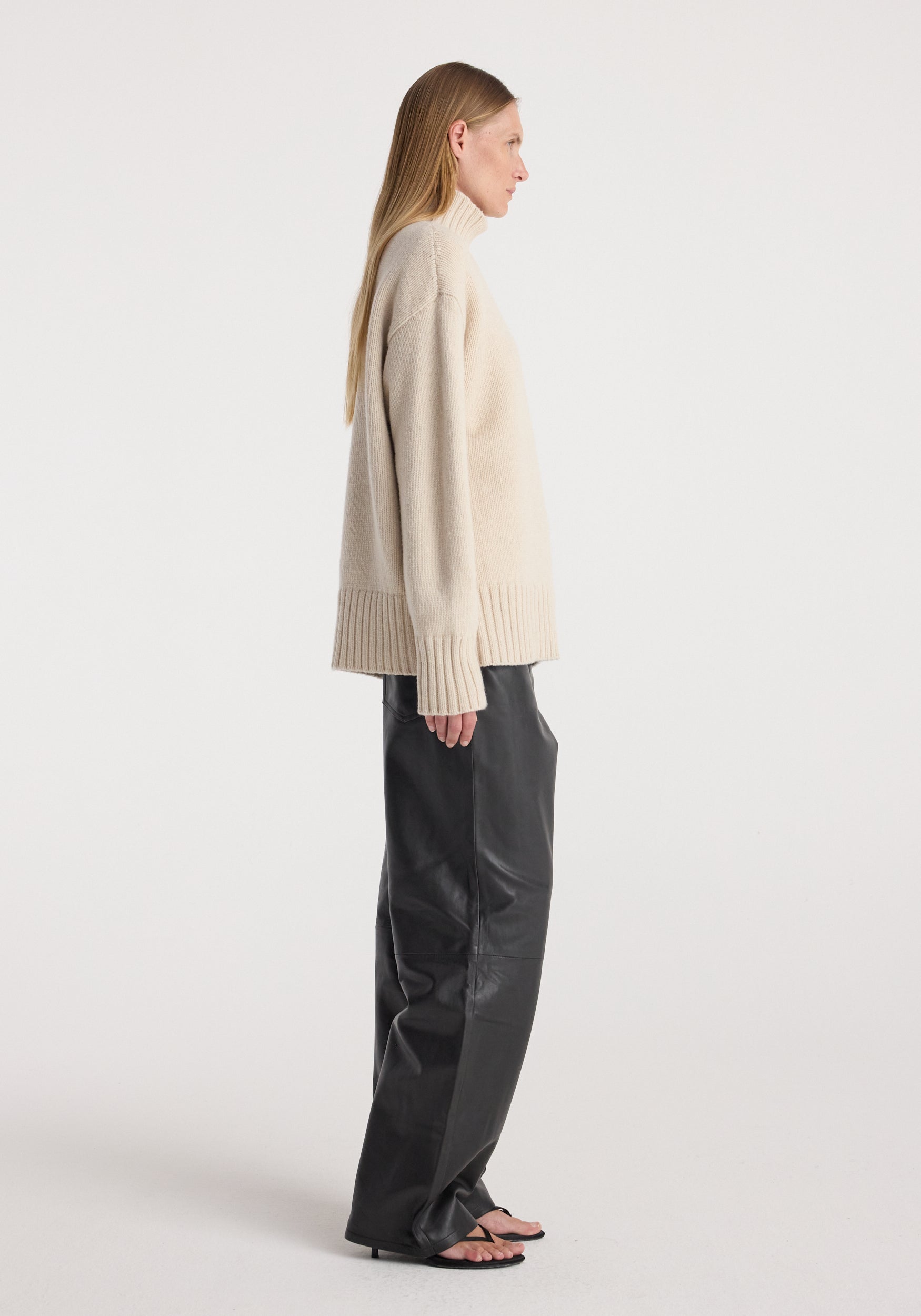 Side view of a woman wearing a Wool-cashmere turtleneck | oat in a soft beige color paired with black wide-leg pants and black open-toe sandals against a plain white background.
