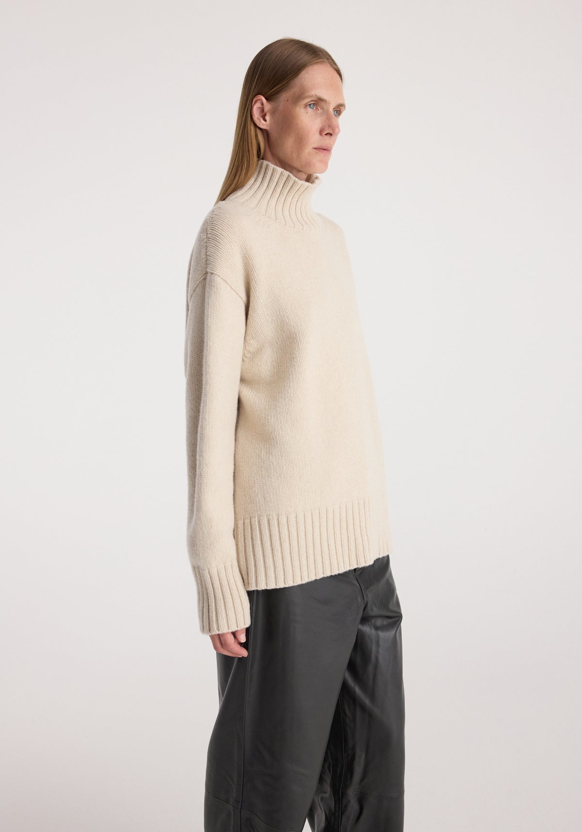 Side view of a female model wearing a soft, cozy Wool-cashmere turtleneck | oat in a relaxed fit, paired with loose black pants against a plain background.