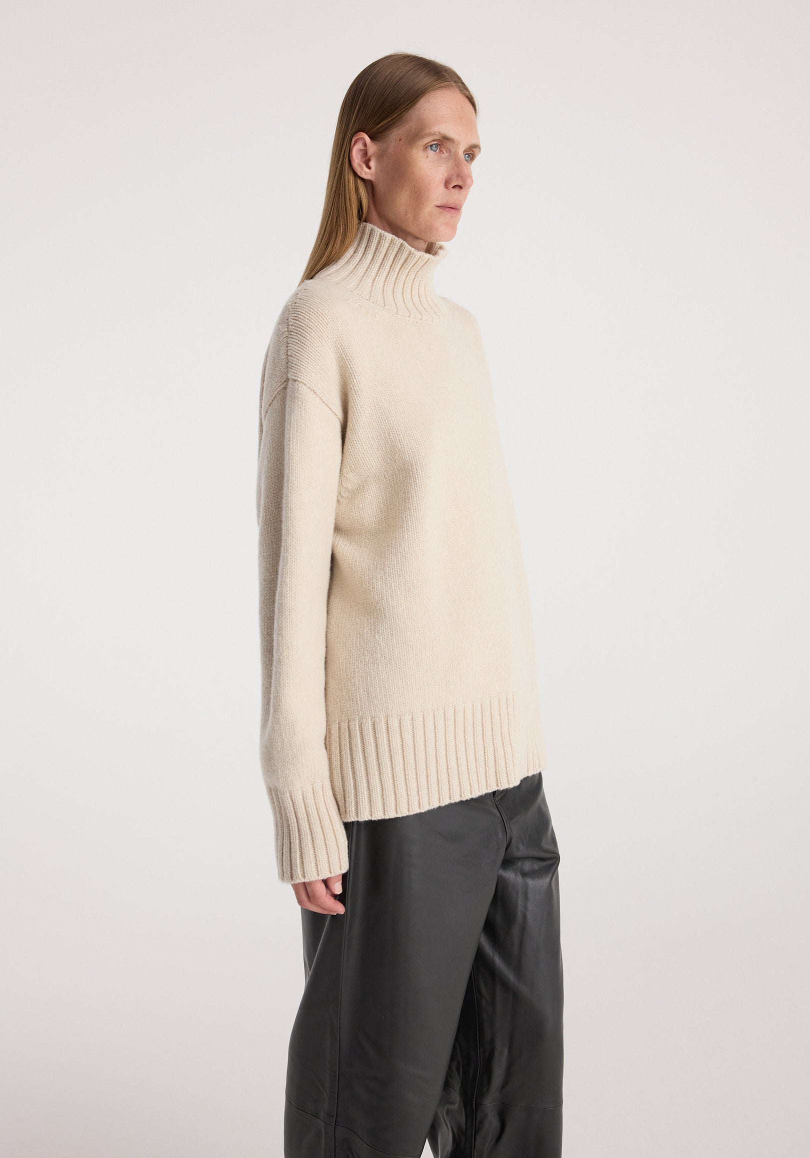 Side view of a female model wearing a soft, cozy Wool-cashmere turtleneck | oat in a relaxed fit, paired with loose black pants against a plain background.
