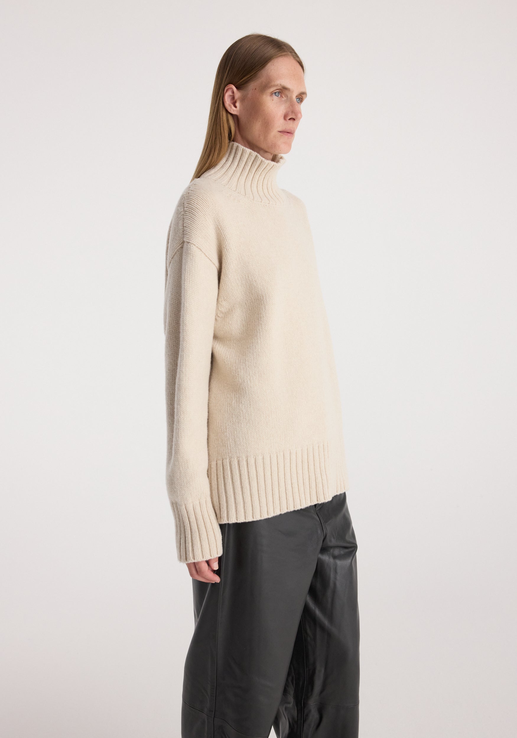 Side view of a female model wearing a soft, cozy Wool-cashmere turtleneck | oat in a relaxed fit, paired with loose black pants against a plain background.