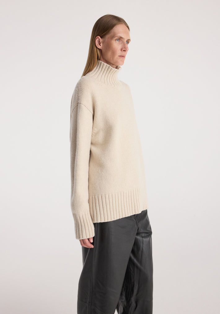 Side view of a female model wearing a soft, cozy Wool-cashmere turtleneck | oat in a relaxed fit, paired with loose black pants against a plain background.