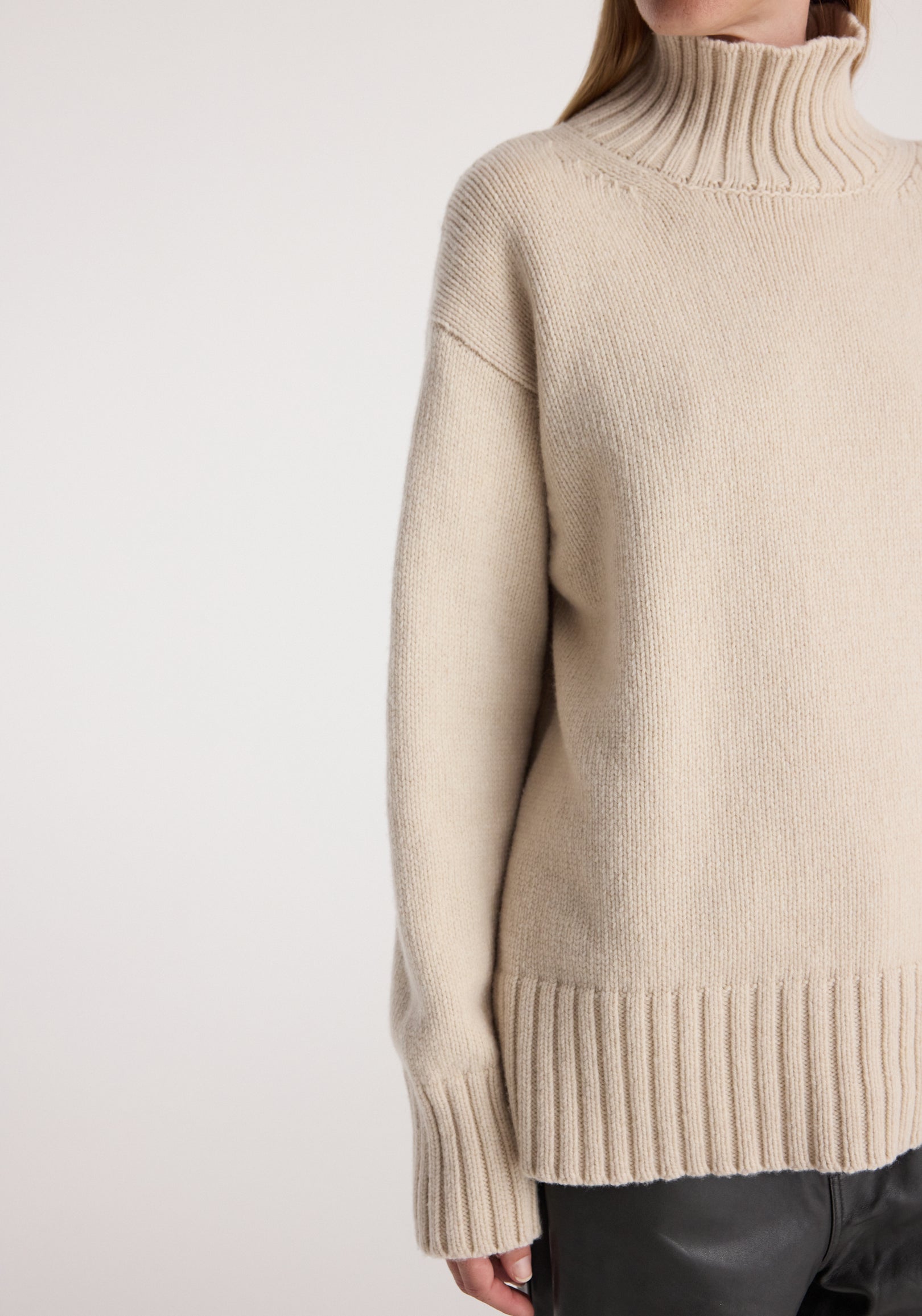 Close-up of a person wearing a soft wool-cashmere turtleneck | oat with ribbed collar, cuffs, and hem, styled with black leather pants against a light background.