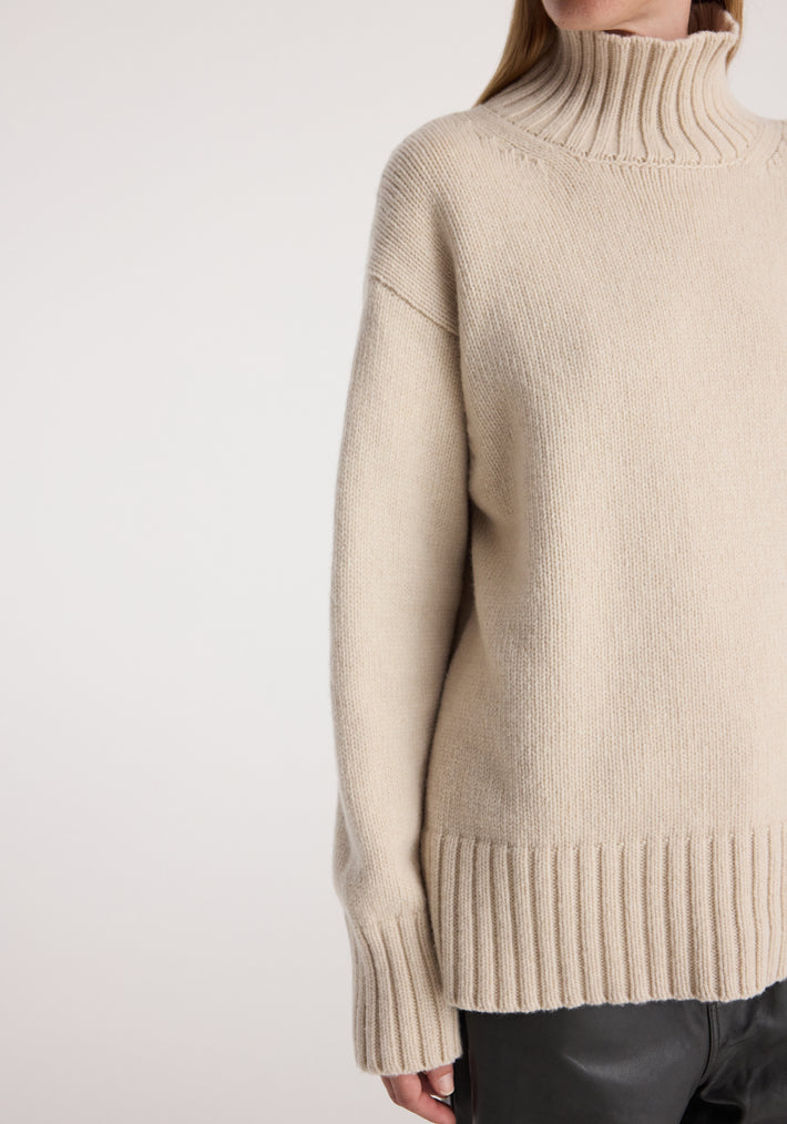 Close-up of a person wearing a soft wool-cashmere turtleneck | oat with ribbed collar, cuffs, and hem, styled with black leather pants against a light background.
