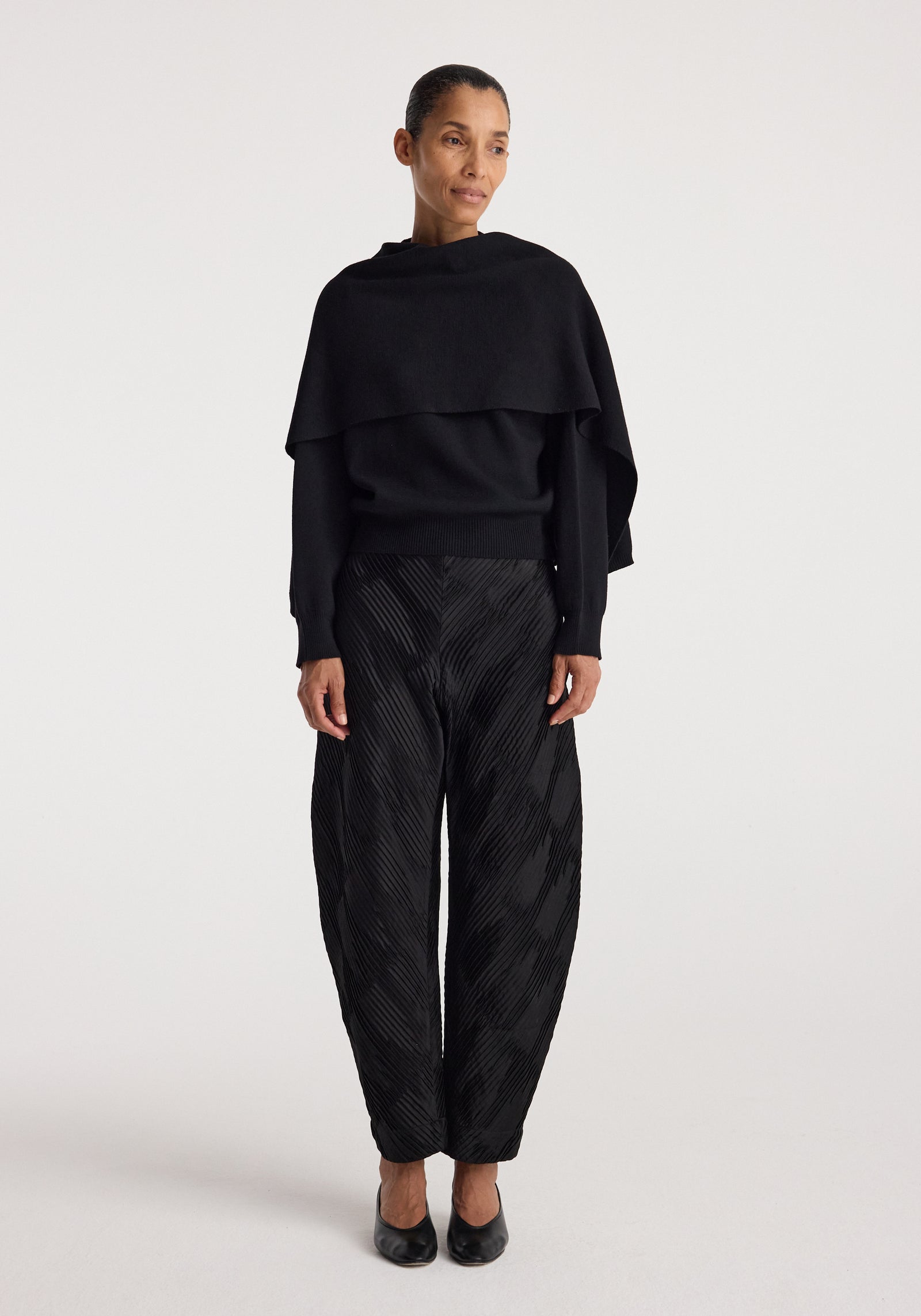 Woman wearing a black layered top with long sleeves and textured black pants, styled with black shoes, showcasing the Cape jumper | black.