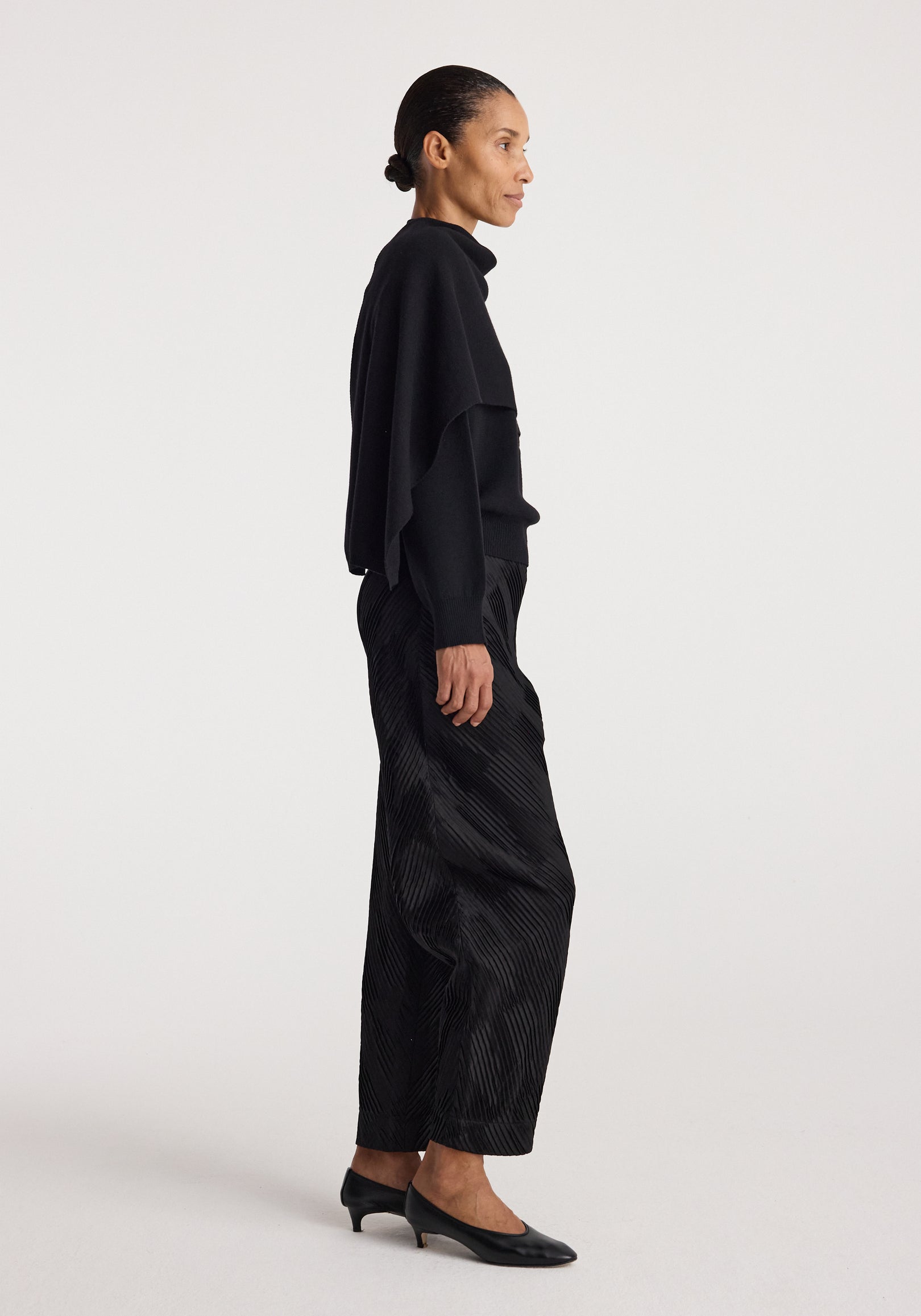 Woman standing in profile wearing a sleek, minimalist black outfit featuring the Cape jumper | black, paired with textured black pants and black low-heeled shoes.