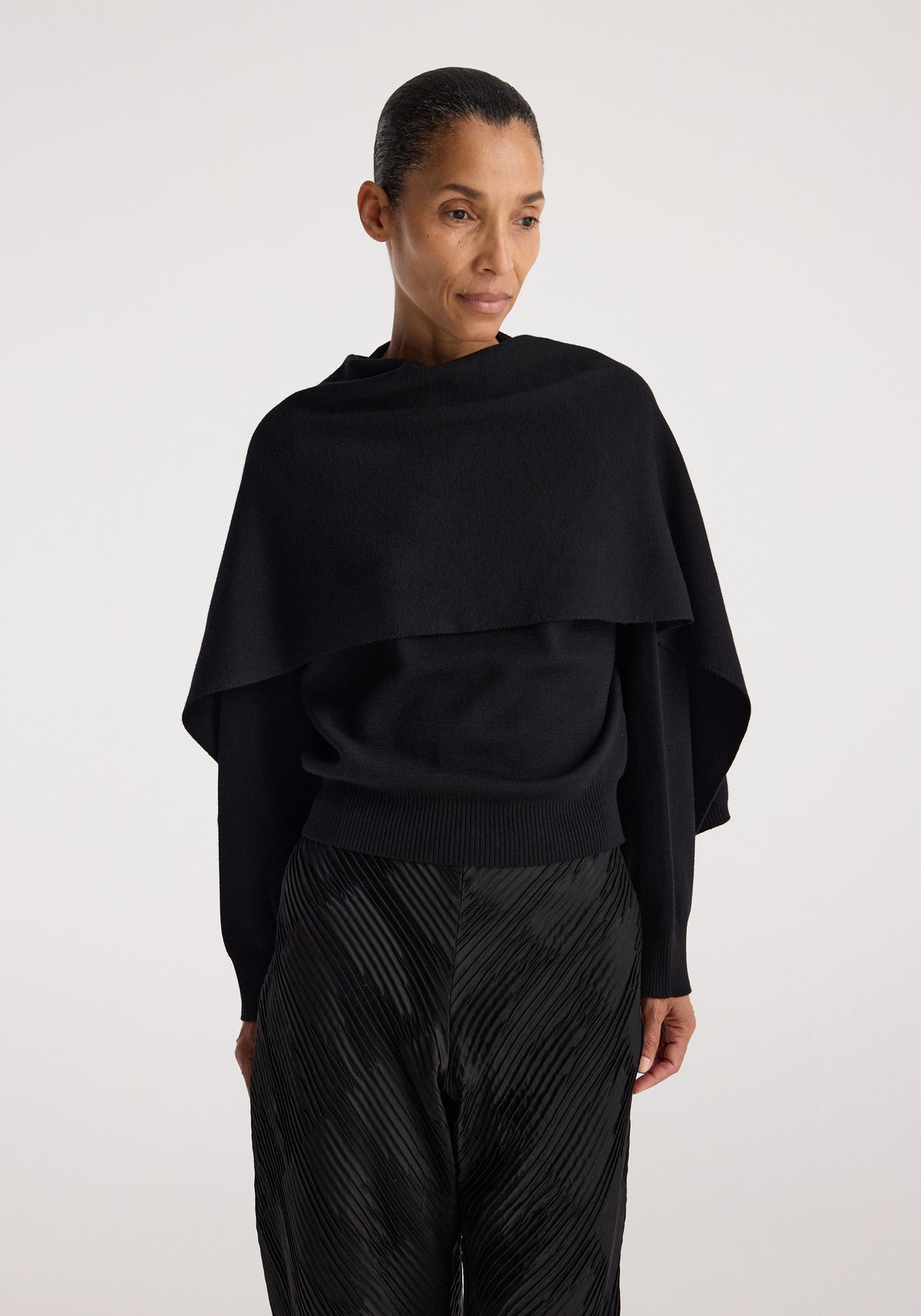 Woman wearing a black Cape jumper | black with a wide neckline and long sleeves, paired with textured black pants, standing against a light background.