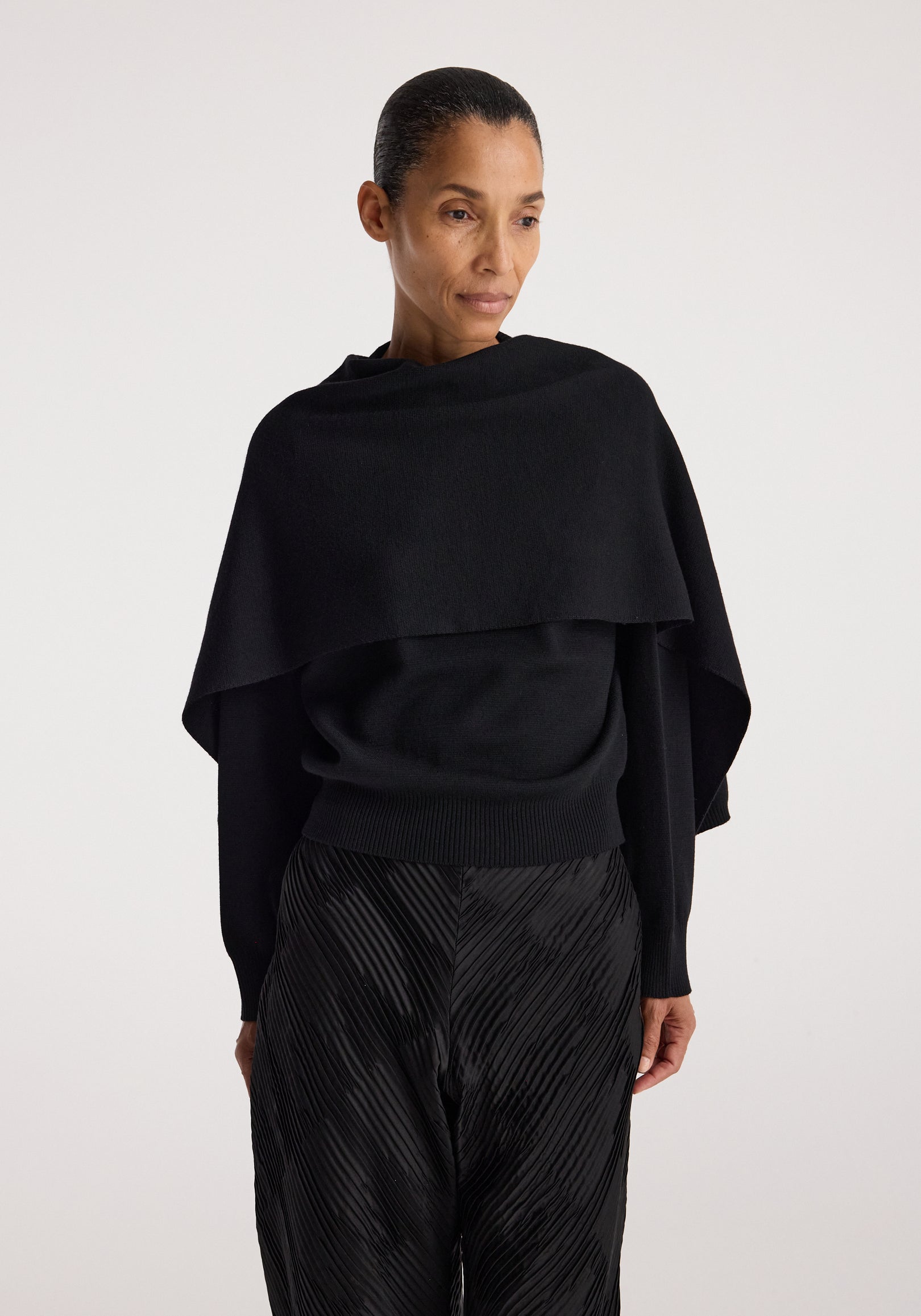 Woman wearing a black Cape jumper | black with a wide neckline and long sleeves, paired with textured black pants, standing against a light background.