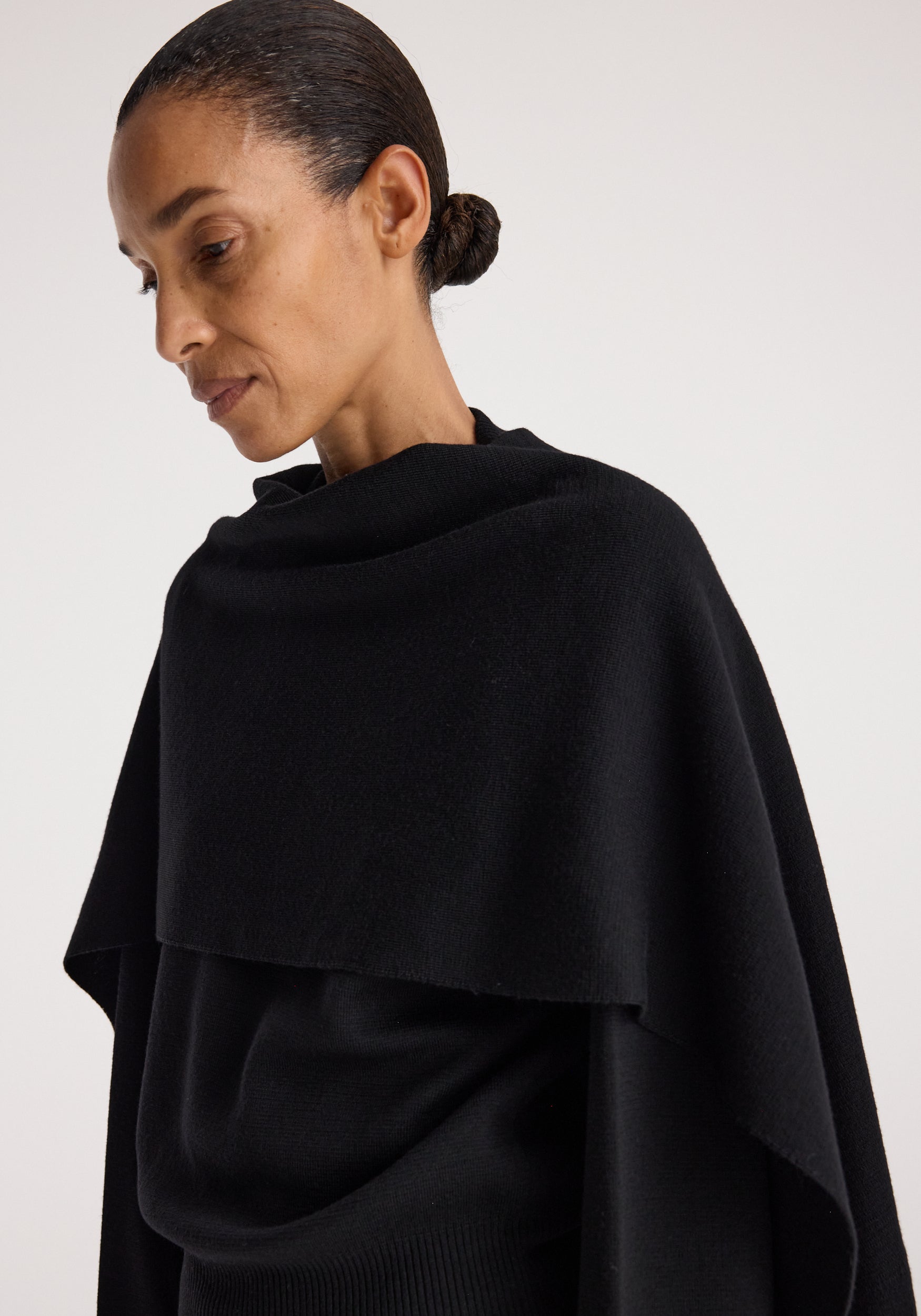 Woman wearing a black Cape jumper | black with a draped neckline and loose fit, styled with hair tied back against a neutral background.
