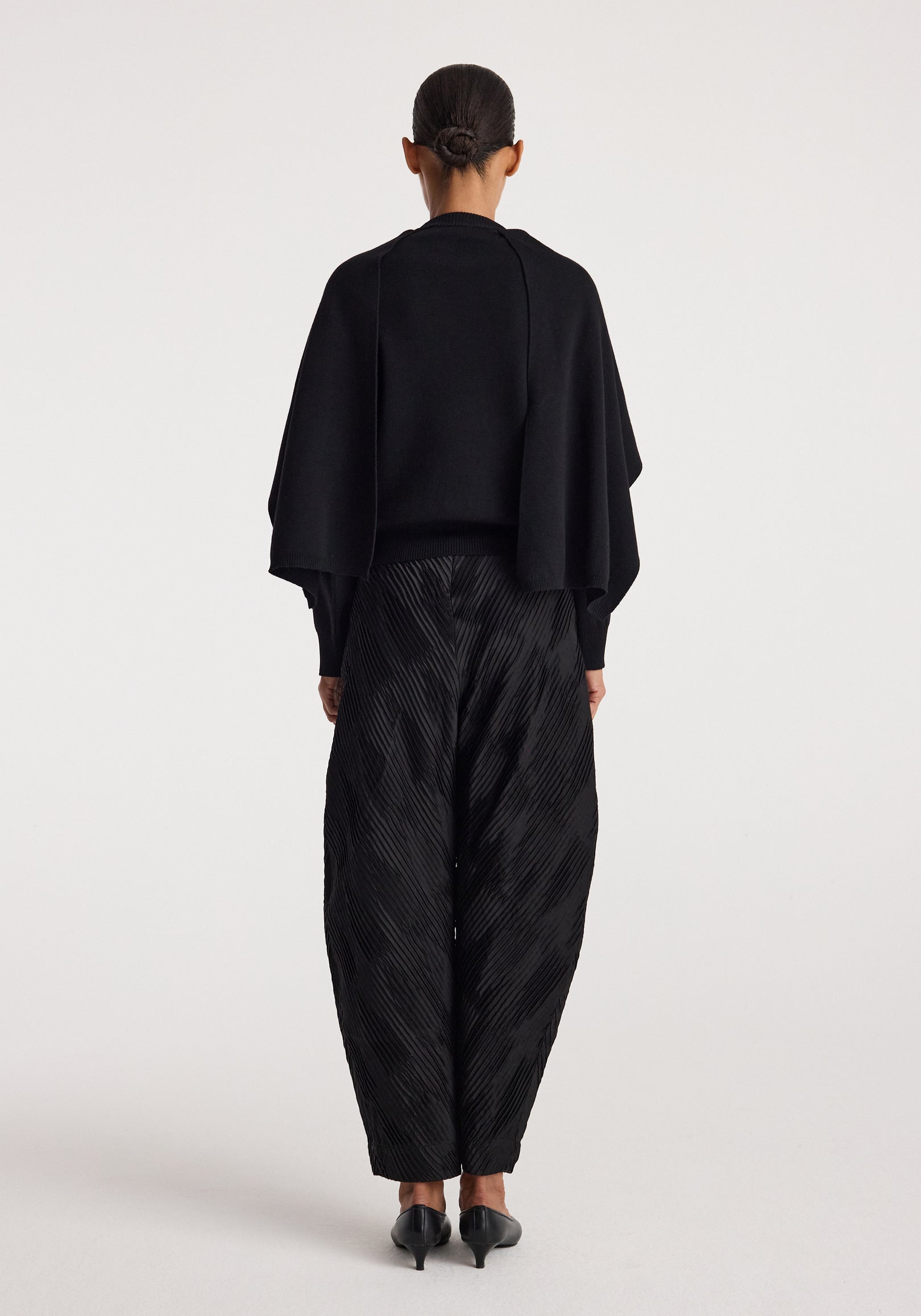 Woman wearing a black cape jumper | black with long sleeves, standing facing away, paired with textured black pants and black heels against a white background