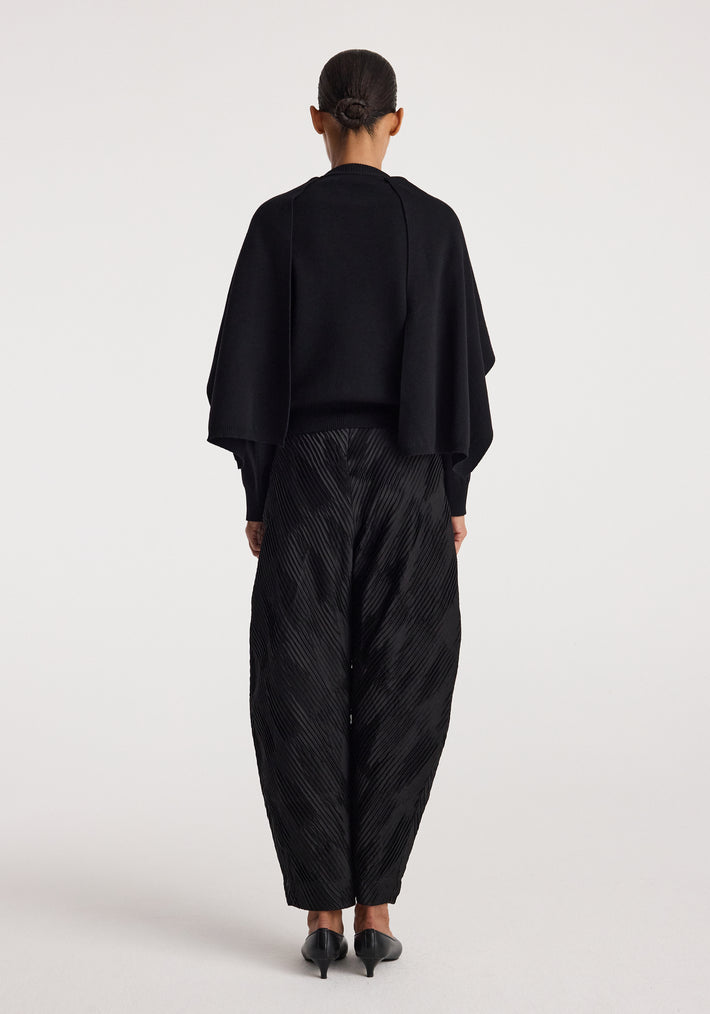 Woman wearing a black cape jumper | black with long sleeves, standing facing away, paired with textured black pants and black heels against a white background