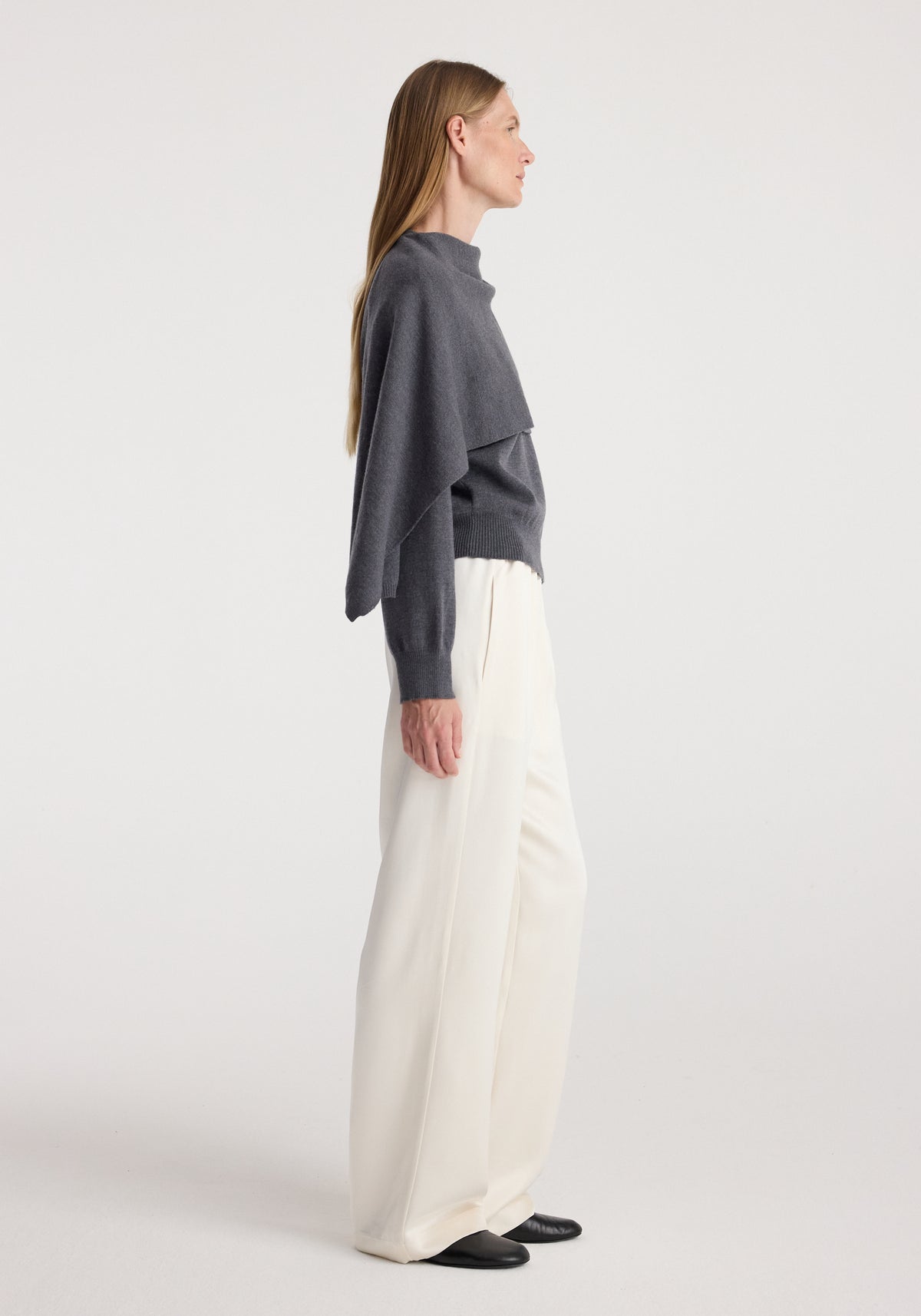 Side view of a woman wearing a dark grey melange cape jumper paired with loose white trousers and black shoes against a plain background.