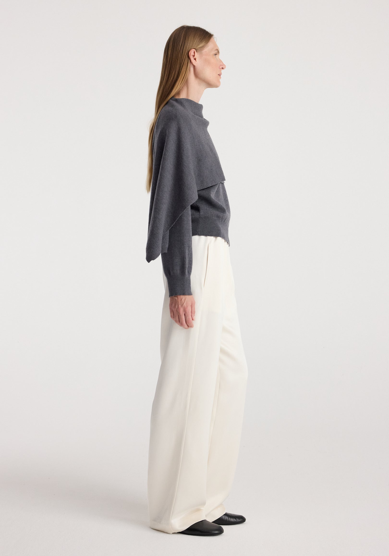Side view of a woman wearing a dark grey melange cape jumper paired with loose white trousers and black shoes against a plain background.
