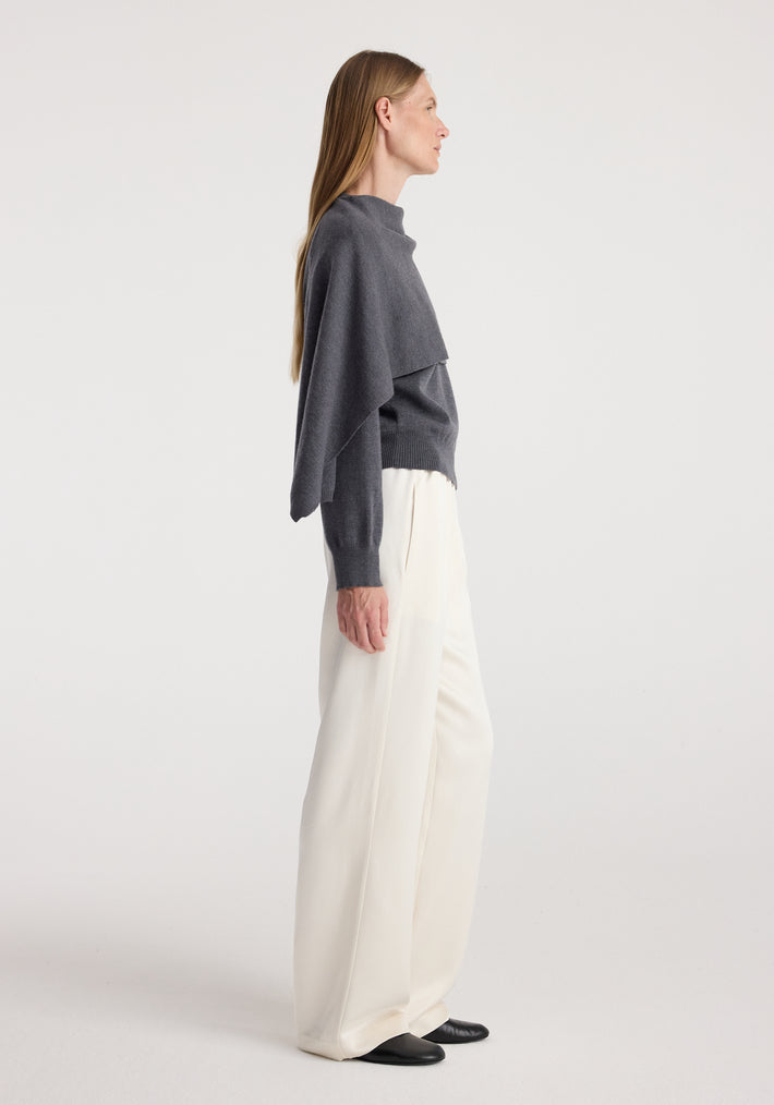 Side view of a woman wearing a dark grey melange cape jumper paired with loose white trousers and black shoes against a plain background.