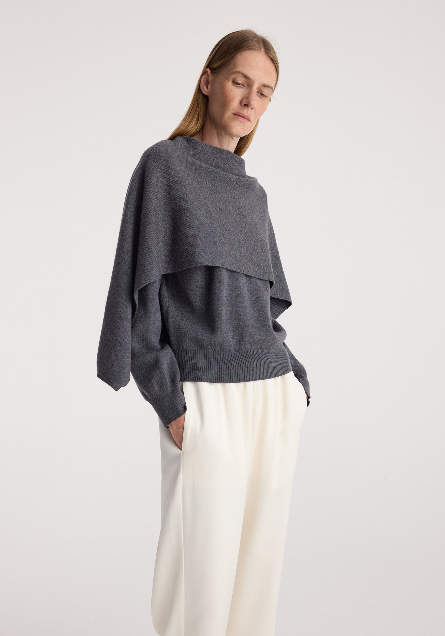 Woman wearing a dark grey melange layered sweater with a high neck and long sleeves paired with loose white trousers, showcasing the Cape jumper | dark grey melange.